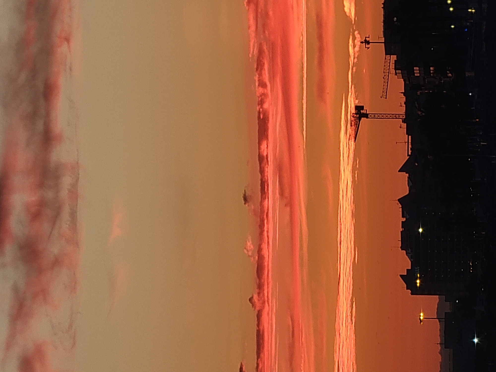 The image captures a vibrant sunset over the city of Quarteira, Portugal. The sky is a dramatic canvas of orange and pink hues, with wispy clouds catching the last rays of sunlight. Silhouetted against this colorful backdrop is the city's skyline, featuring several buildings, some appearing to be under construction with cranes visible. One prominent building has lights on, illuminating some of its windows. A street lamp with a bright light also stands out. The overall scene is a picturesque blend of natural beauty and urban development.