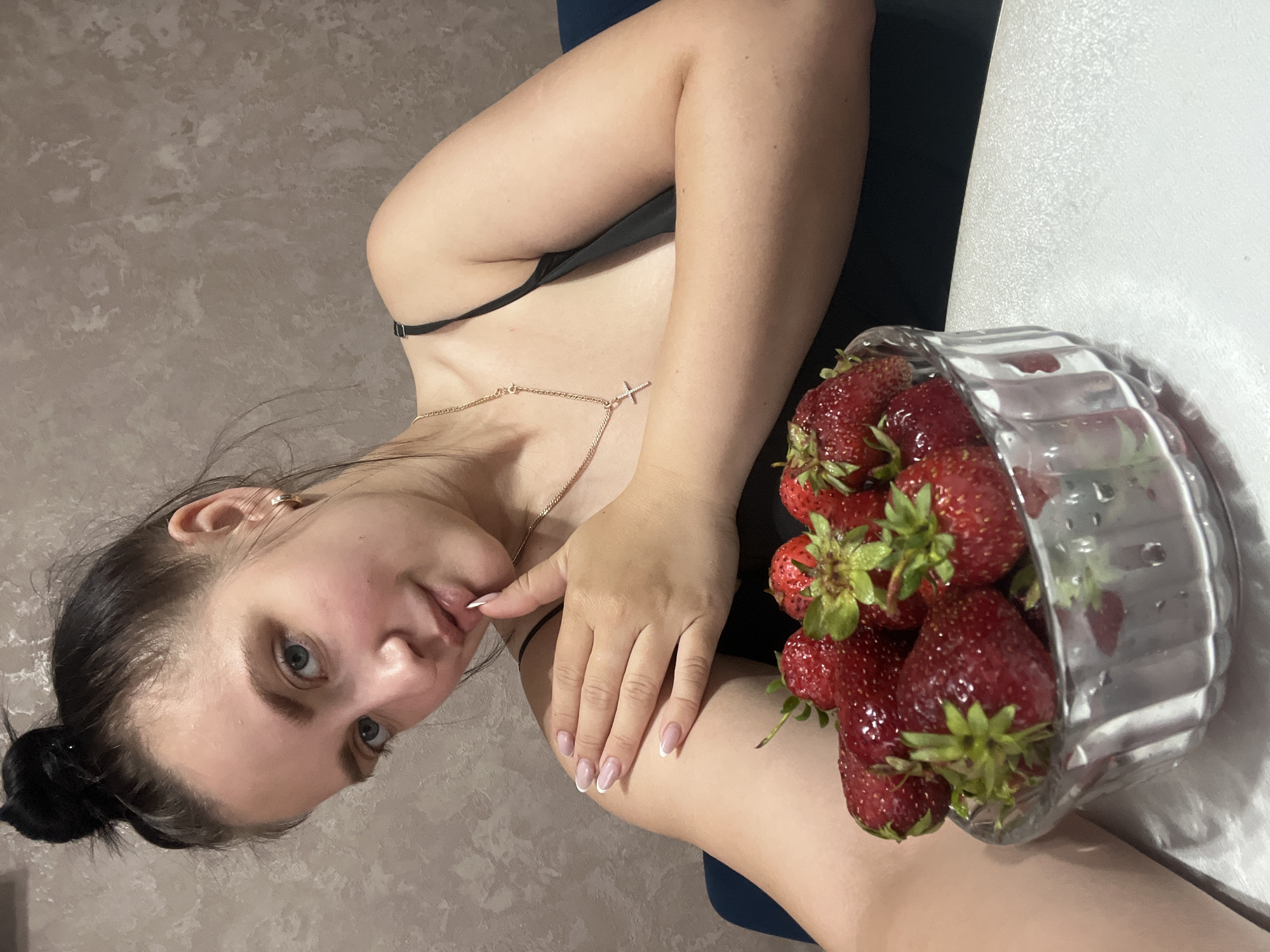 A young woman with dark hair pulled into a bun poses with a bowl of strawberries. She is wearing a black camisole and a gold necklace with a cross pendant. Her left arm is resting on her shoulder, and her hand is near her mouth. The background is a textured wall. The scene appears to be indoors, possibly in a residential setting in Марковский, Russia.