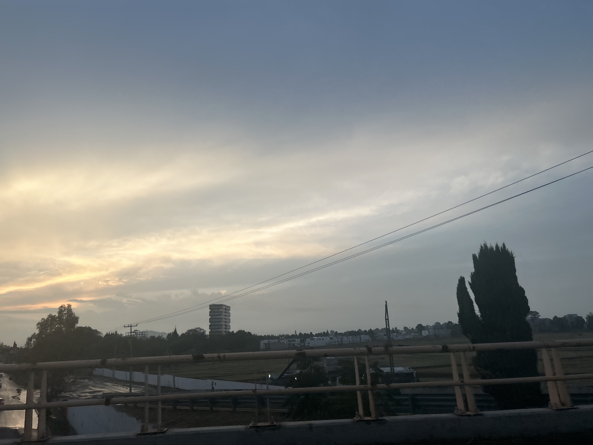 The image depicts an urban landscape at dawn or dusk, possibly in the city of Unknown, Unknown. The sky is a mix of light blues and yellows, suggesting either sunrise or sunset. Overhead power lines stretch across the upper portion of the frame.
In the midground, a bridge railing is visible, suggesting the image was taken from a bridge. Behind the railing, there is a landscape featuring trees, buildings, and what appears to be a tall, monolithic structure that may be an unfinished or unique building. A dense, dark tree stands on the right side of the frame.
The foreground consists primarily of the bridge structure itself, rendered in muted tones. The overall scene has a hazy, somewhat muted quality, potentially due to weather conditions or atmospheric effects.
