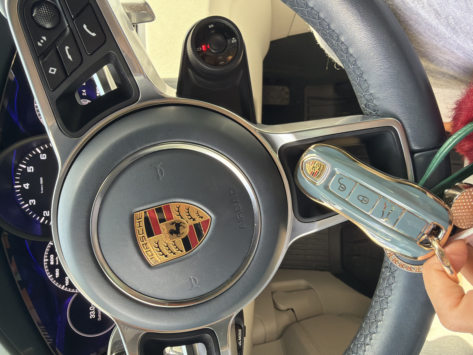 A close-up, eye-level shot shows a person's hand holding a light blue Porsche car key with gold trim and a diamond-encrusted keyring. The key is positioned in front of a dark gray leather steering wheel with metallic accents. The Porsche logo is visible in the center of the steering wheel. The dashboard displays various gauges, including a speedometer reading up to 200 km/h and a digital display showing "33.0" and "Outside." To the right of the steering wheel, buttons for media and phone controls are visible, along with a small digital display reading "2.4". A rotary dial with settings "0", "I", "S", and "S+" is also present. The interior of the car appears to be light-colored. The scene suggests the car is parked or in a low-speed environment. The image was taken inside a vehicle, and no external environment is visible. The specific location in Tbilisi, Georgia, cannot be determined from the visual information provided.