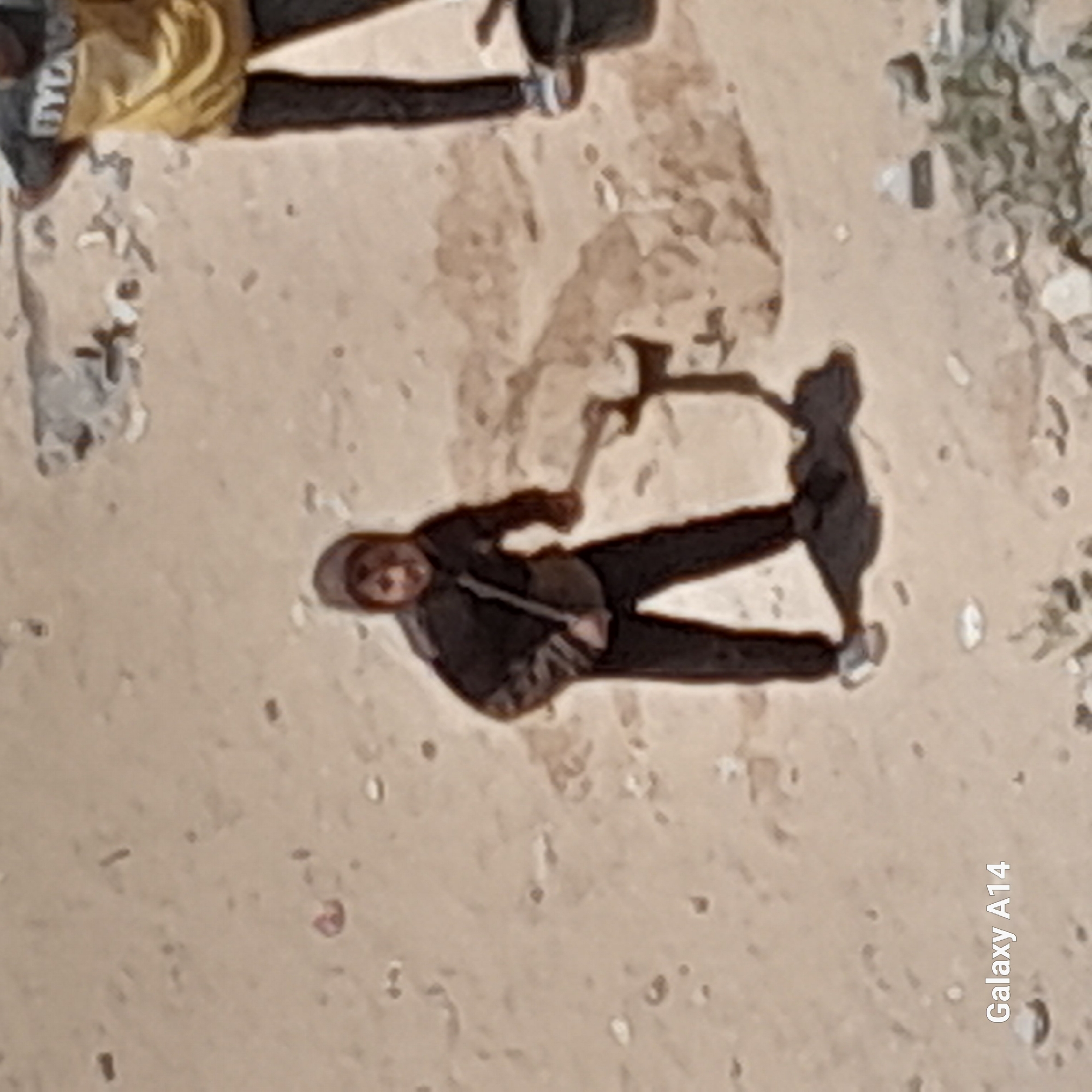 Here's a detailed description of the image:
**Scene Overview:**
The image appears to be taken from an elevated, possibly aerial perspective, looking down on a desert-like or sandy environment. The lighting suggests it may be daytime with the sun casting shadows from figures.
**People:**
- The image contains at least two people. One person is visible at the bottom center, wearing dark clothing (possibly pants and a jacket) and a head covering. The other person is located at the top, and is wearing light color clothing ( possibly pants and a jacket).
- The posture of both people suggests they are in a state of walking.
**Objects:**
- A long handled object, possibly a tool or equipment, is seen by the side of the person at the bottom.
**Environment:**
- The surface appears to be arid and dry, likely a desert environment, characterized by sandy or earthen texture.
- Patches of vegetation or debris are scattered across the scene.
**Miscellaneous:**
- The text "Galaxy A14" is visible in the bottom right corner. This indicates the image was likely taken with a Samsung Galaxy A14 smartphone.