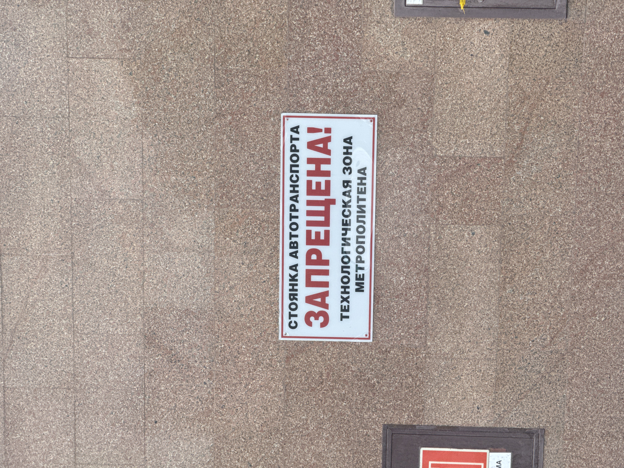 The image presents a top-down view of a light brown, speckled paved surface, likely granite or similar stone, composed of large rectangular tiles with subtle grout lines. Dominating the center is a white rectangular sign with a red border, positioned vertically.
The sign displays a clear prohibition in Russian text. The first line reads "СТОЯНКА АВТОТРАНСПОРТА" (Parking of motor vehicles). Below this, in large, bold red capital letters, is "ЗАПРЕЩЕНА!" (FORBIDDEN!). Further down, the sign specifies the reason for the prohibition: "ТЕХНОЛОГИЧЕСКАЯ ЗОНА" (TECHNOLOGICAL ZONE) and "МЕТРОПОЛИТЕНА" (OF THE METROPOLITAN / METRO). This indicates that the area is a restricted technical zone belonging to the metro system, and therefore vehicle parking is not allowed.
The lighting is bright and even, suggesting it is daytime. No people or vehicles are visible in the scene, focusing attention on the regulatory sign. In the bottom right corner, a small, dark rectangular object is partially visible, appearing to be part of another sign or utility cover, with only indistinct letters like "MA" visible. Given the text and the provided context, the location is Saint Petersburg, Russia, likely near a metro station or an associated service area.