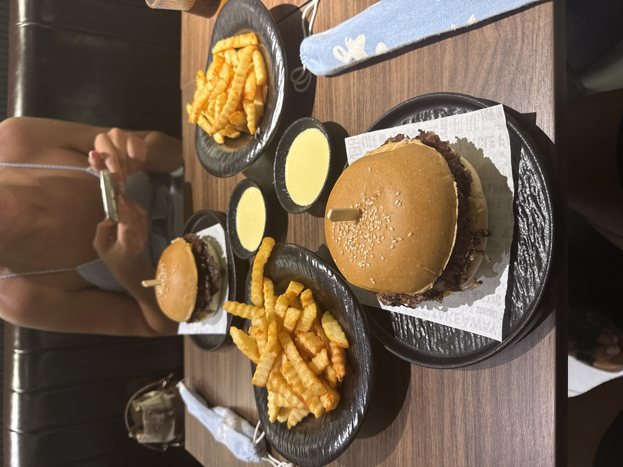 A table at a restaurant shows two hamburgers on black plates and two plates of French fries, also on black plates, plus two small bowls of yellow sauce. One person is seen holding a mobile phone, and the scene appears to be in Đà Nẵng, Vietnam.
