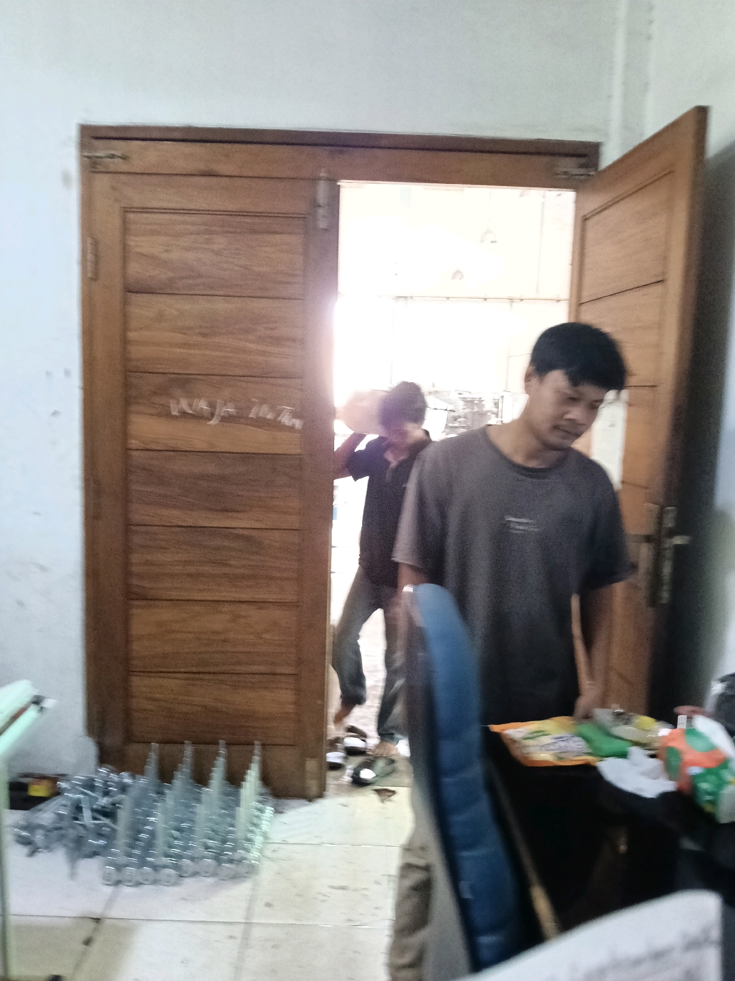 In a room with a white ceiling and walls, a wooden double door stands slightly ajar. Sunlight streams in from the opening, illuminating the scene. On the left, a portion of a table with a light green surface is visible. In front of the door, a collection of metal components is neatly arranged on the floor.
Two men are present. The man on the right is in the foreground, facing towards the left and slightly downwards. He is wearing a dark grey t-shirt and appears to be looking at something on a desk to his right. The desk has a dark surface and is cluttered with various items, including a bag of snacks and a white paper. A blue chair is partially visible in front of the desk.
The second man is in the background, on the left side of the doorway, partially obscured by the open door. He is wearing a dark shirt and light-colored pants and is walking towards the right, away from the camera. He is carrying a light-colored object over his shoulder.
The environment suggests an indoor setting, possibly a workshop or storage area. The presence of metal components and the general appearance of the room do not provide specific clues to indicate the location within the city of Belimbing, Indonesia.