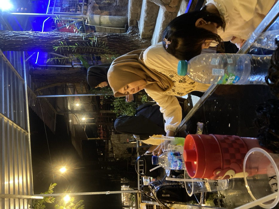 The scene depicts a nighttime gathering at an outdoor or semi-outdoor dining establishment in Cembor, Indonesia. Four individuals are visible, seated at a dark, reflective table, with evidence of at least one more person out of frame.

In the center, a woman wearing a tan hijab and a white, long-sleeved top with dark embroidered patterns is seated, holding a clear plastic cup containing a green liquid and a straw. Behind her, partially obscured, is another woman in a black hijab and dark clothing. Further back and to the right, a third woman in a black hijab is partially visible. To the right of the central woman, a young girl with dark hair and a white top is seated, looking down towards the table. A small light-colored hair clip is present in the girl's hair. Dark, curly hair in the bottom foreground indicates another seated individual.

On the table in the foreground are multiple items: a prominent red insulated cup with a checkered pattern and a character emblem, several clear plastic cups (some with straws), one "Aqua" brand bottled water, and one "Cleo's" brand bottled water. A black quilted shoulder bag with a metal chain strap is visible resting on the table behind the red cup.

The environment is illuminated by various artificial light sources, including string lights with warm yellow bulbs, overhead lighting, and blue accent lights on structures in the background. An overhead corrugated metal roof structure is partially visible. In the background, multiple wooden and metal structures indicate a multi-level establishment, with large tree trunks integrated into the architecture on the right. Stone or concrete steps lead upwards on the far right. Sparse foliage is present throughout the scene.