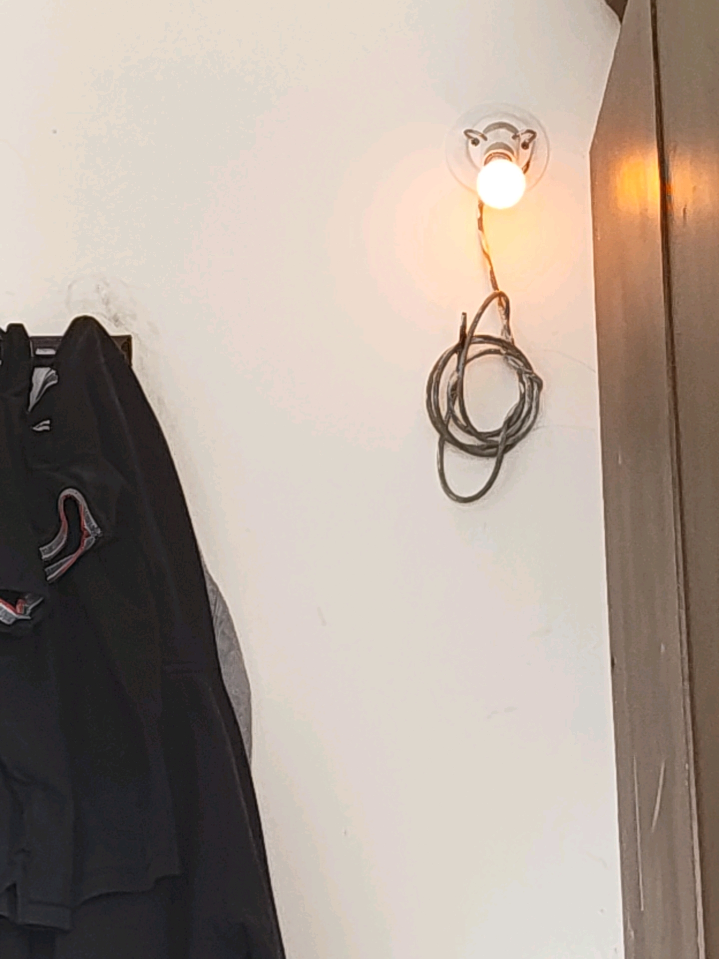 A close-up shot, likely indoors, shows a simple ceiling light fixture with a single, illuminated bulb. A coiled black wire hangs loosely from the fixture, suspended against a plain, off-white wall. To the left, a portion of a dark garment, possibly a jacket or coat, hangs on a rack or hook. The garment is dark, with red and white striped accents visible on the fabric. To the right, a section of a wooden surface, likely a door or cabinet, is visible, appearing to be a muted brown color with some vertical scratch marks. The lighting suggests an interior space, possibly a hallway or entryway, with the warm glow of the bulb creating subtle shadows. There are no people visible in the image. The location is not specified in the image itself but based on the prompt, it's suggested to be in Tathawade, India.
