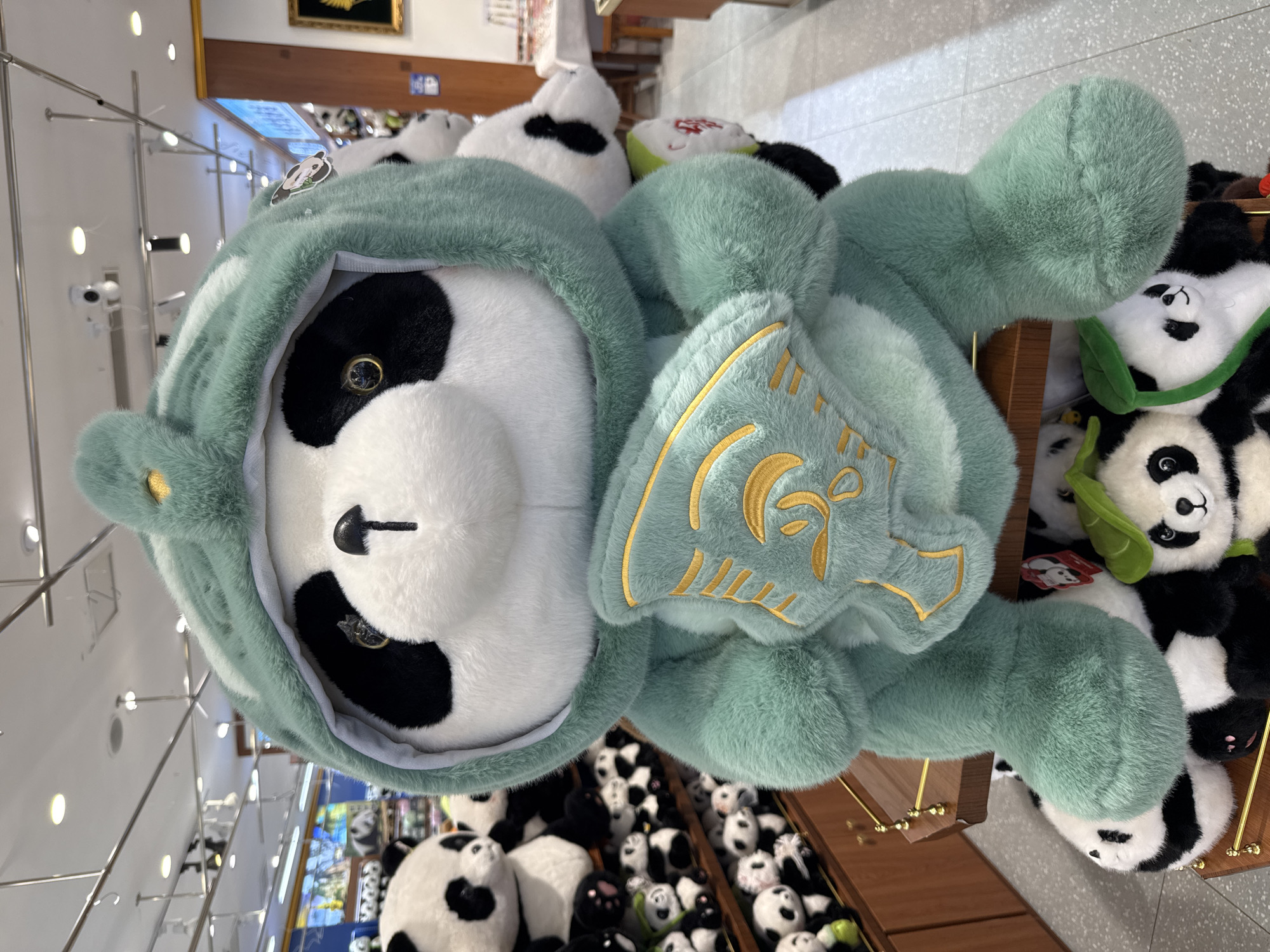 The image shows a close-up of a plush panda toy wearing a light green hoodie with an emblem. The panda is primarily white with black patches around its eyes and small black button nose. It appears to be lying on top of or among other plush panda toys. The location, Jinjiang District in China, is inferred from the details in the image such as product choice and store aesthetics. A rack filled with assorted merchandise is also visible. The scene implies a commercial environment, likely a retail store or souvenir shop.