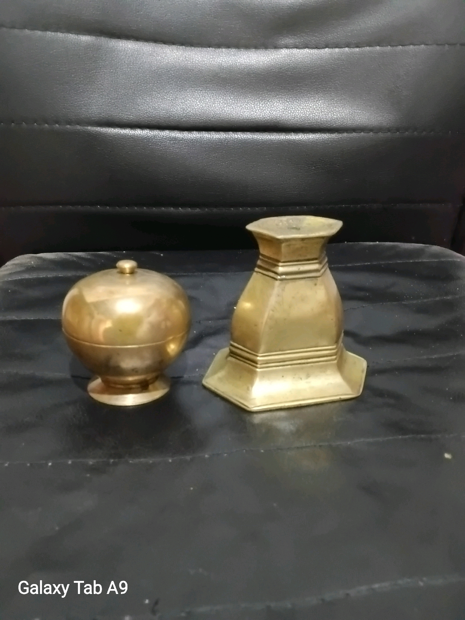 Two antique brass objects are displayed on a dark, textured surface, possibly a table or chair. The object on the left is spherical with a small knob on top and rests on a circular base. The object on the right has a hexagonal opening at the top and a wider, stepped base. The background consists of a dark, ribbed material, likely leather. The lighting is dim, casting subtle reflections on the brass. The overall impression is one of stillness and historical preservation. This image does not provide any information about the city of Karangan, Indonesia, or its specific location within the city.