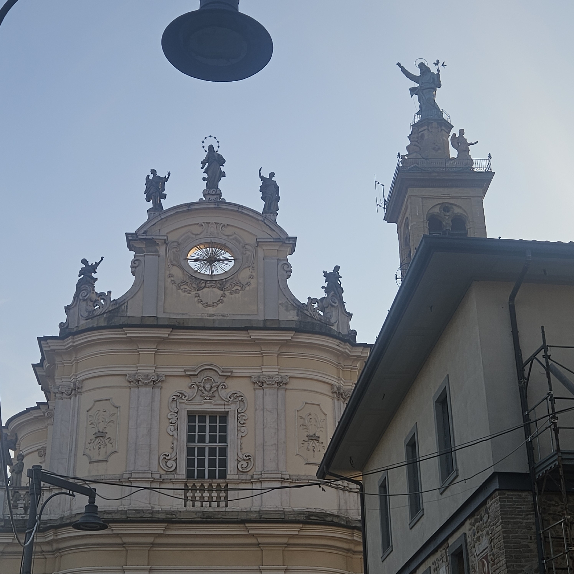 In the city of Calcinate, Italy, an image captures a partial view of the Chiesa di Santa Maria Assunta, also known as the Duomo di Calcinate, a prominent church. The facade of the church, characterized by its baroque architectural style, is adorned with statues of saints and intricate decorative elements. A large, dark lamppost is prominently positioned in the foreground, partially obscuring the sky. To the right of the church, another building, lighter in color and with a simpler facade, is visible, partially obscuring the church's tower and bell. The bell tower, a slender structure, is topped with a statue of a figure with outstretched arms, possibly a saint or religious icon, holding what appears to be birds. The sky is clear and a pale blue, suggesting daylight. The overall scene depicts a street view with religious architecture and urban elements.
