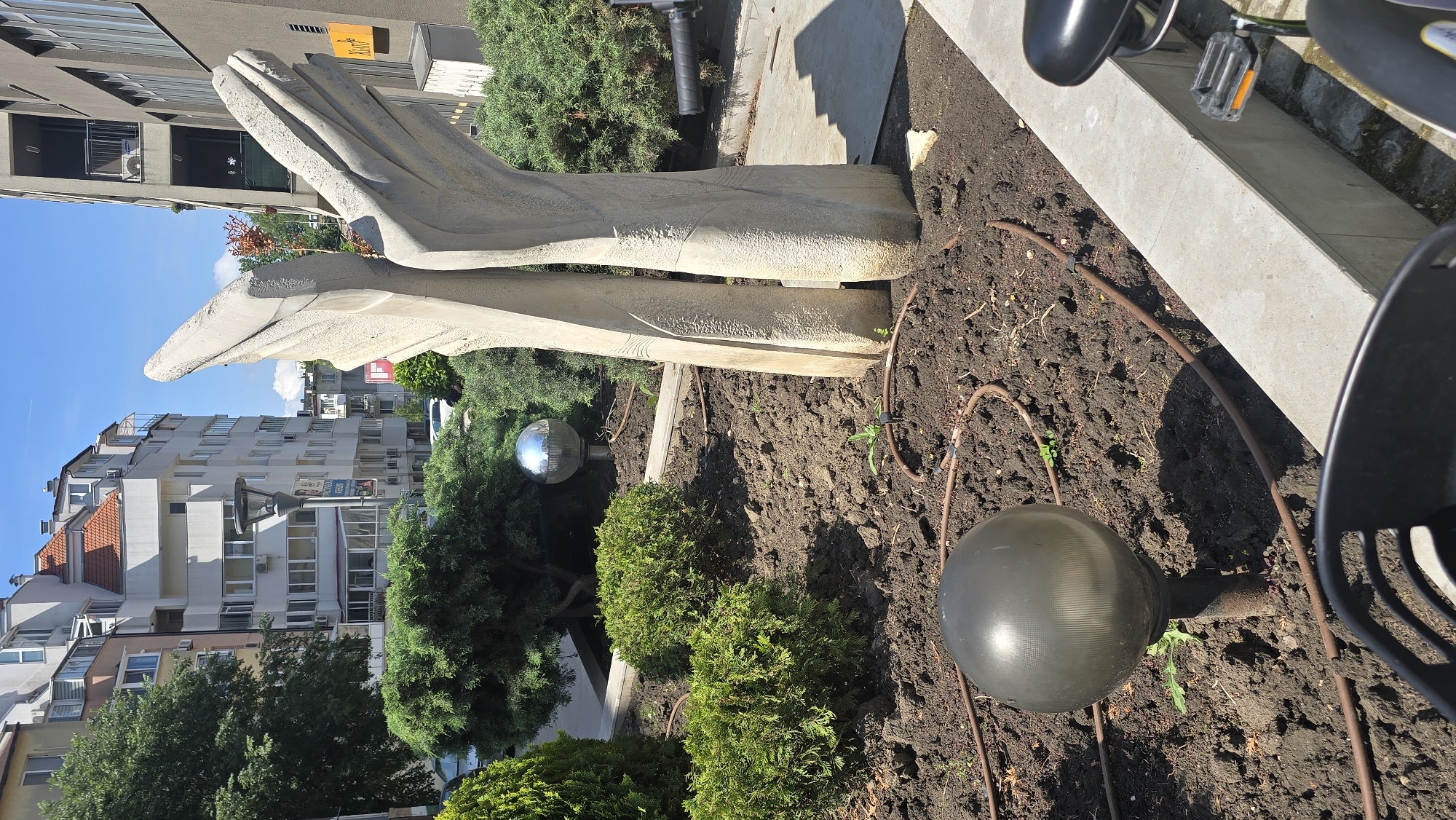 The image shows a sculpture in a garden bed in Varna, Bulgaria. The sculpture is light-colored and abstract, with two elongated, wing-like shapes. The garden bed contains soil, small evergreen bushes, and spherical outdoor lights. A brown irrigation hose is visible on the ground. Buildings are visible in the background, including residential structures. A bicycle with a black seat and pedals is partially visible on the right side of the image. The sky is blue with some wisps of clouds.