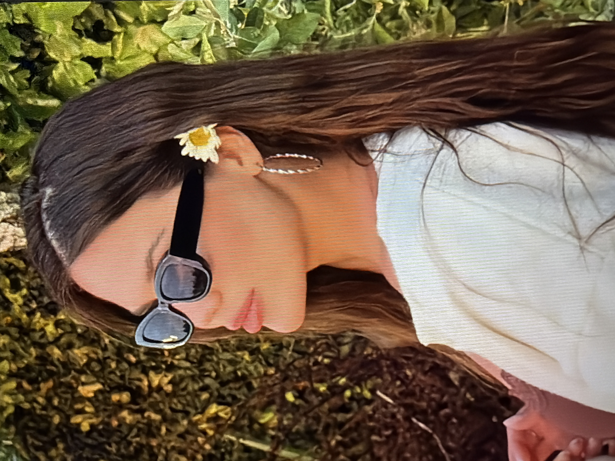 In a medium shot, a woman with long, dark brown hair is seen from the side, facing left. She is wearing black, rectangular sunglasses and a white top. A white daisy with a yellow center is tucked behind her right ear. She also has a large, twisted hoop earring in her right ear. The background consists of lush green foliage, suggesting an outdoor setting. No specific location in Sirius, Russia, can be identified from the image.