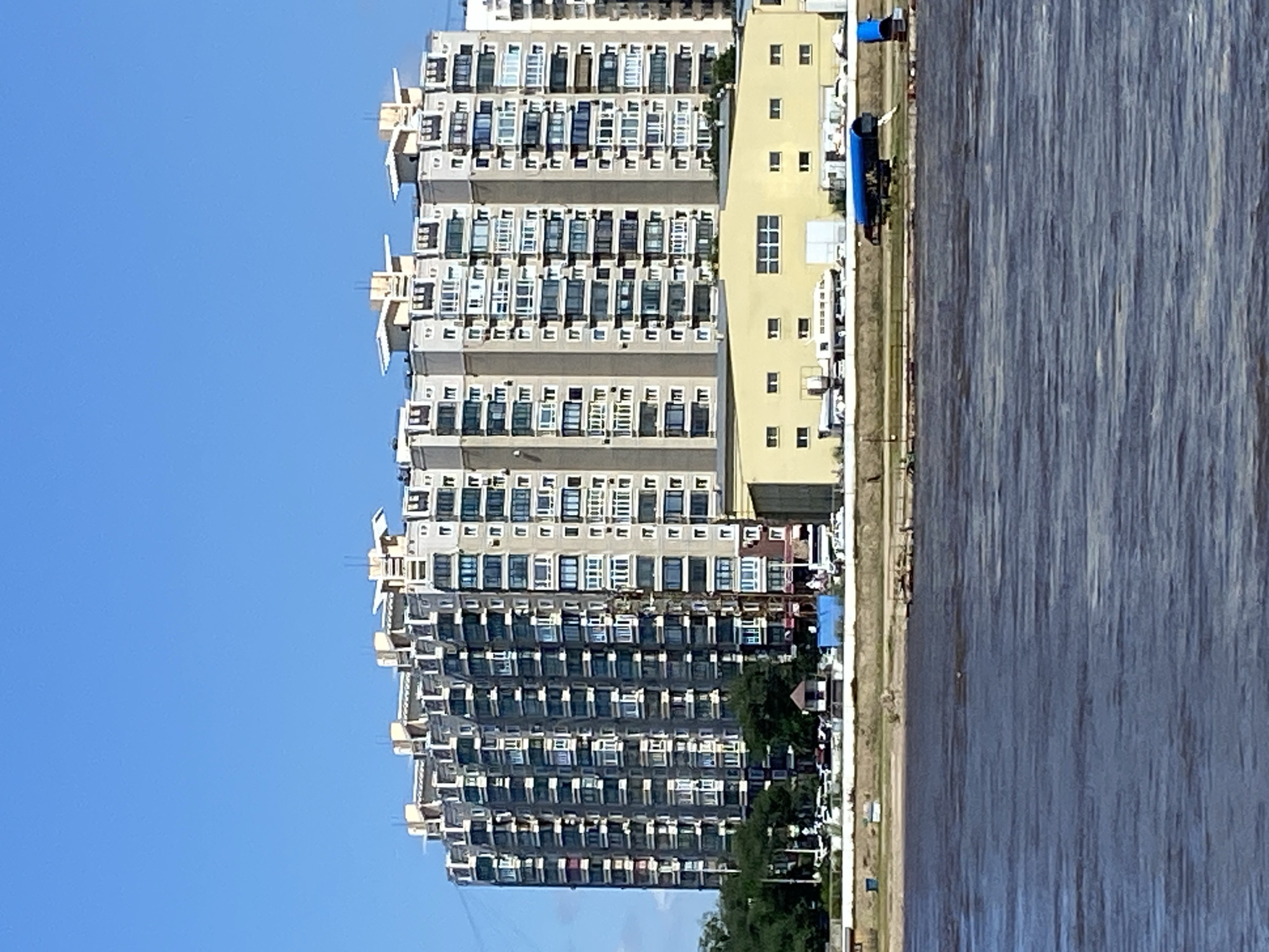 The image depicts a view of a large, multi-story apartment complex situated along a riverfront in Blagoveshchensk, Russia. The building, a prominent feature, is light-colored with numerous windows and balconies, suggesting a residential or mixed-use purpose. In the foreground, a body of dark, choppy water, likely the Amur River, dominates the lower half of the frame. The riverbank area behind the water features a strip of land with some greenery and a low-rise yellow building. Several vehicles are parked near this building, and what appears to be a small dock or pier extends into the water on the right side. The sky is clear and blue, indicating fair weather. The overall scene conveys an urban waterfront landscape.