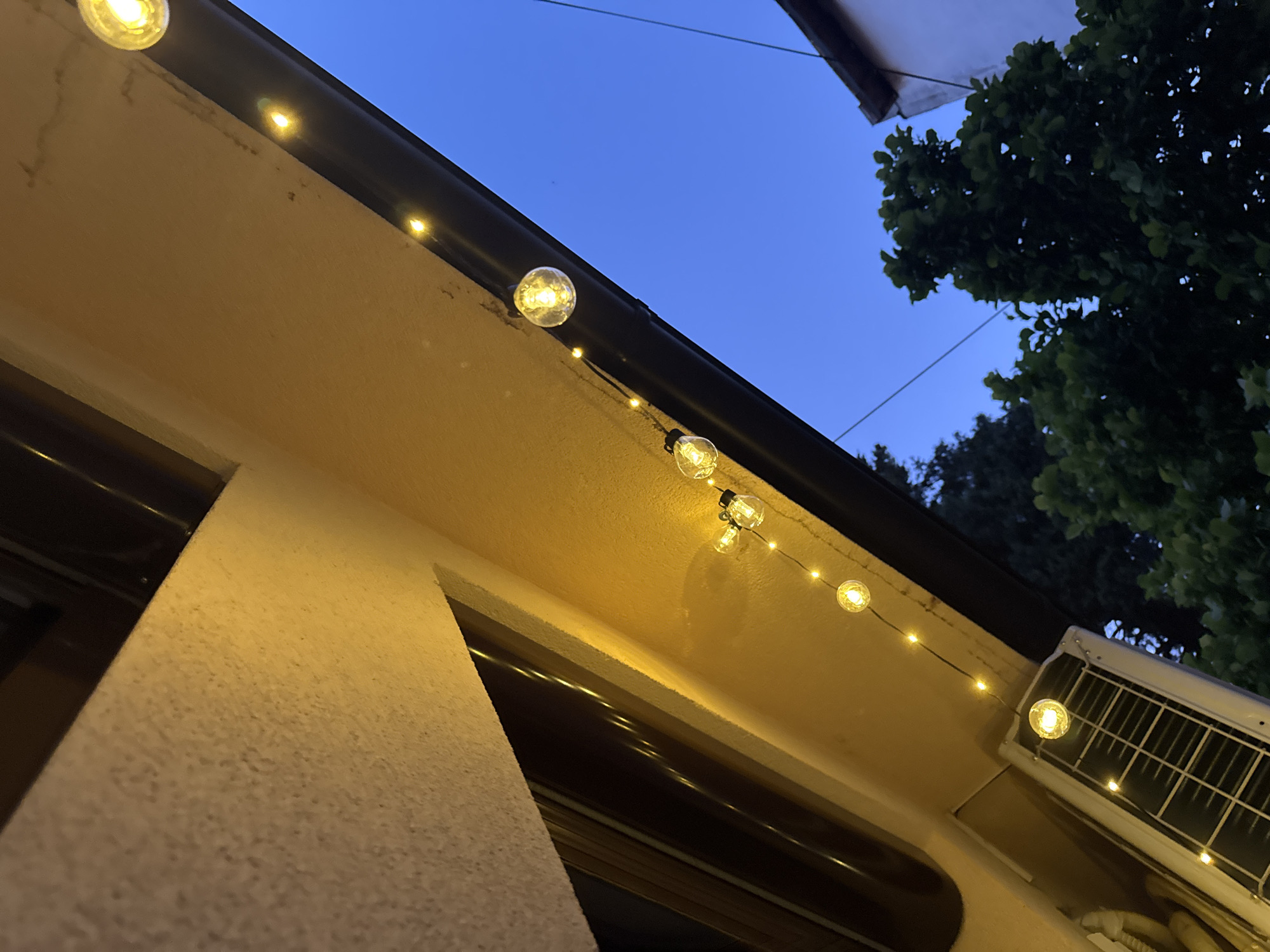 The image shows a string of yellow fairy lights hanging from an eave on the exterior of a building, likely in Zvezdara Urban Municipality, Serbia. The lights are illuminated, casting a warm glow. Below the eave, an outdoor air conditioning unit is visible. The sky is a deep blue, indicating it is twilight or early evening. Trees and a portion of another building are also visible in the background. The perspective is looking upwards, from a low angle.