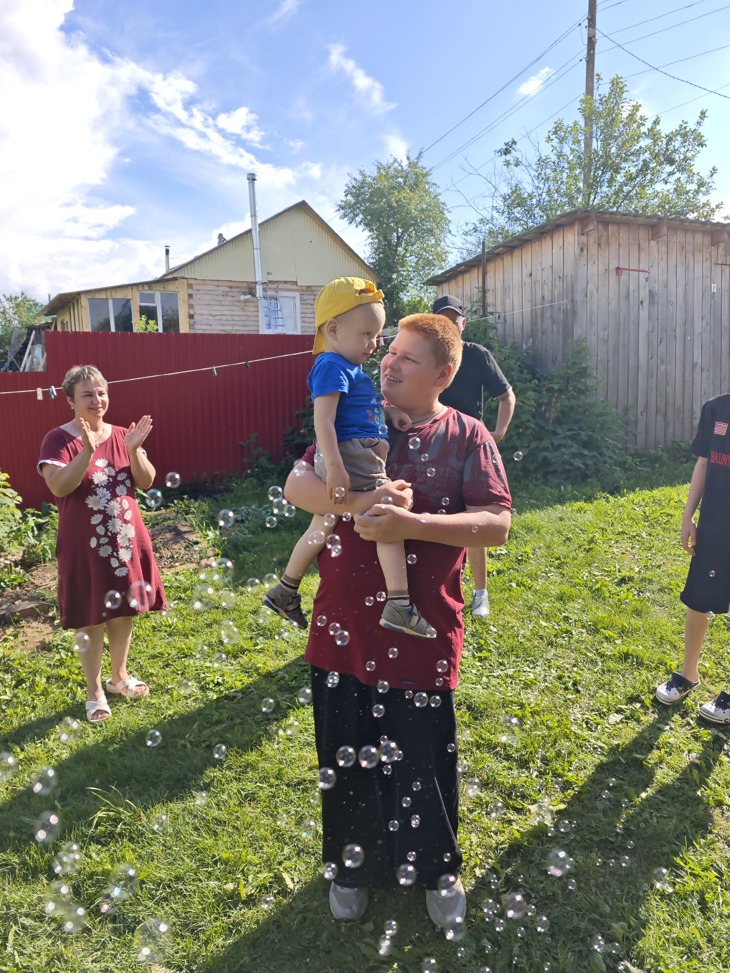 In a sunny outdoor setting, likely a backyard in Фоки, Russia, a group of people are enjoying a playful moment. The scene is illuminated by bright daylight, suggesting it is daytime. Lush green grass covers the ground, with several wooden structures, including a house and a shed, visible in the background. A red fence runs across the mid-ground, adding a splash of color.
The central focus is a young boy wearing a blue t-shirt and a yellow baseball cap, being held by a teenage boy with red hair. The older boy is dressed in a maroon t-shirt and black pants. They are looking at each other, with the older boy smiling.
To the left, a woman in a burgundy dress with a floral pattern is clapping her hands, her face lit up with a smile, indicating amusement or approval. Further in the background, another person wearing a black t-shirt and a cap is visible. To the far right, partially visible, is another individual in a black t-shirt and shorts.
The area is filled with numerous floating soap bubbles, catching the sunlight and creating a whimsical atmosphere. The presence of bubbles suggests an activity like bubble blowing, contributing to a lighthearted and joyful event. The overall impression is one of family fun and a pleasant summer day. No visible text is present in the image.