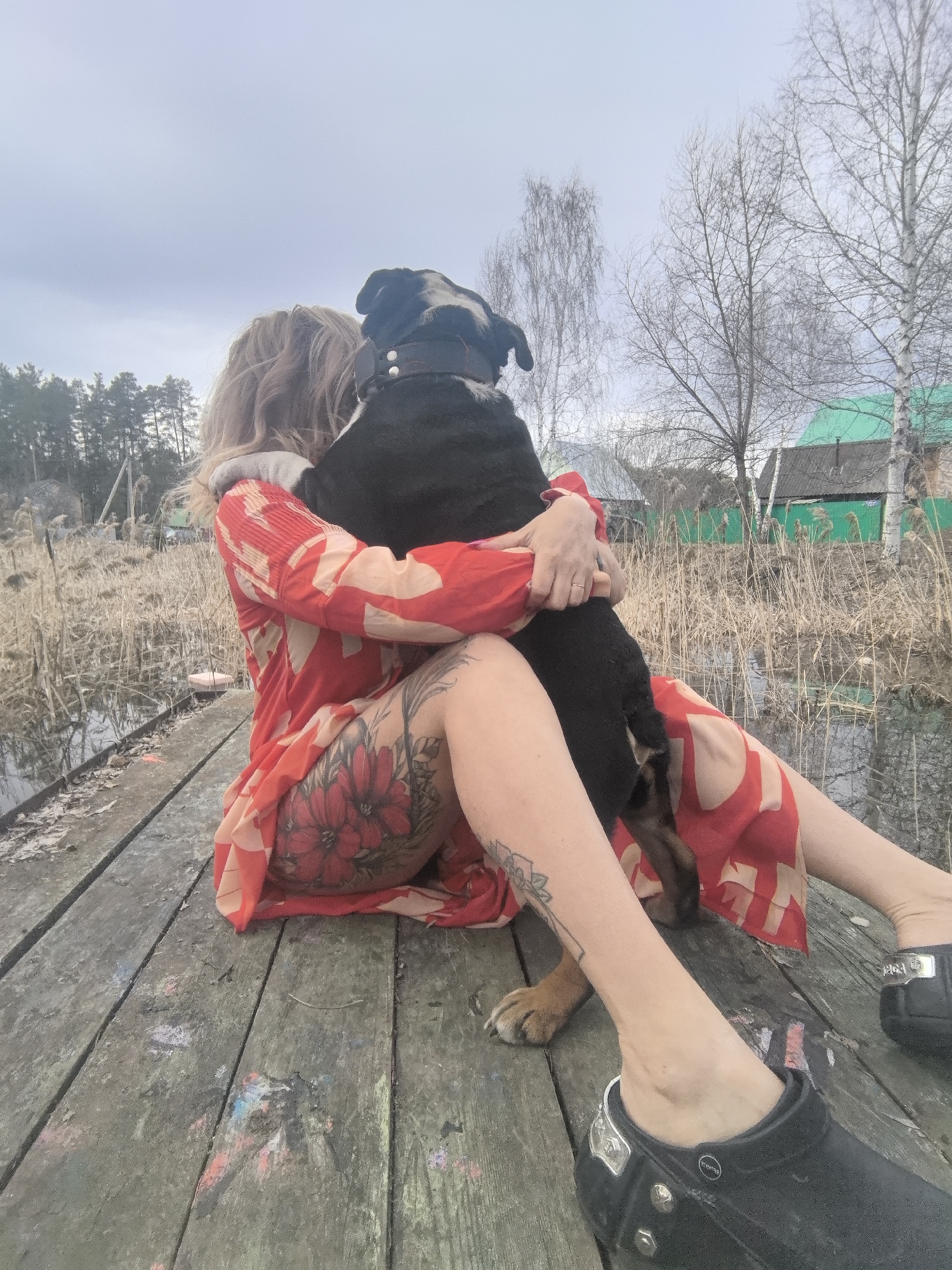 A person is seated on a wooden dock, embracing a black dog. The person is wearing a red and white patterned dress and black clogs. A large floral tattoo is visible on the person's thigh. The dog is wearing a black collar. The background shows a body of water, reeds, trees, and a building with a green roof and green siding. The setting appears to be rural or semi-rural. The weather appears cloudy. The scene is located in the city of Noviy, Russia.