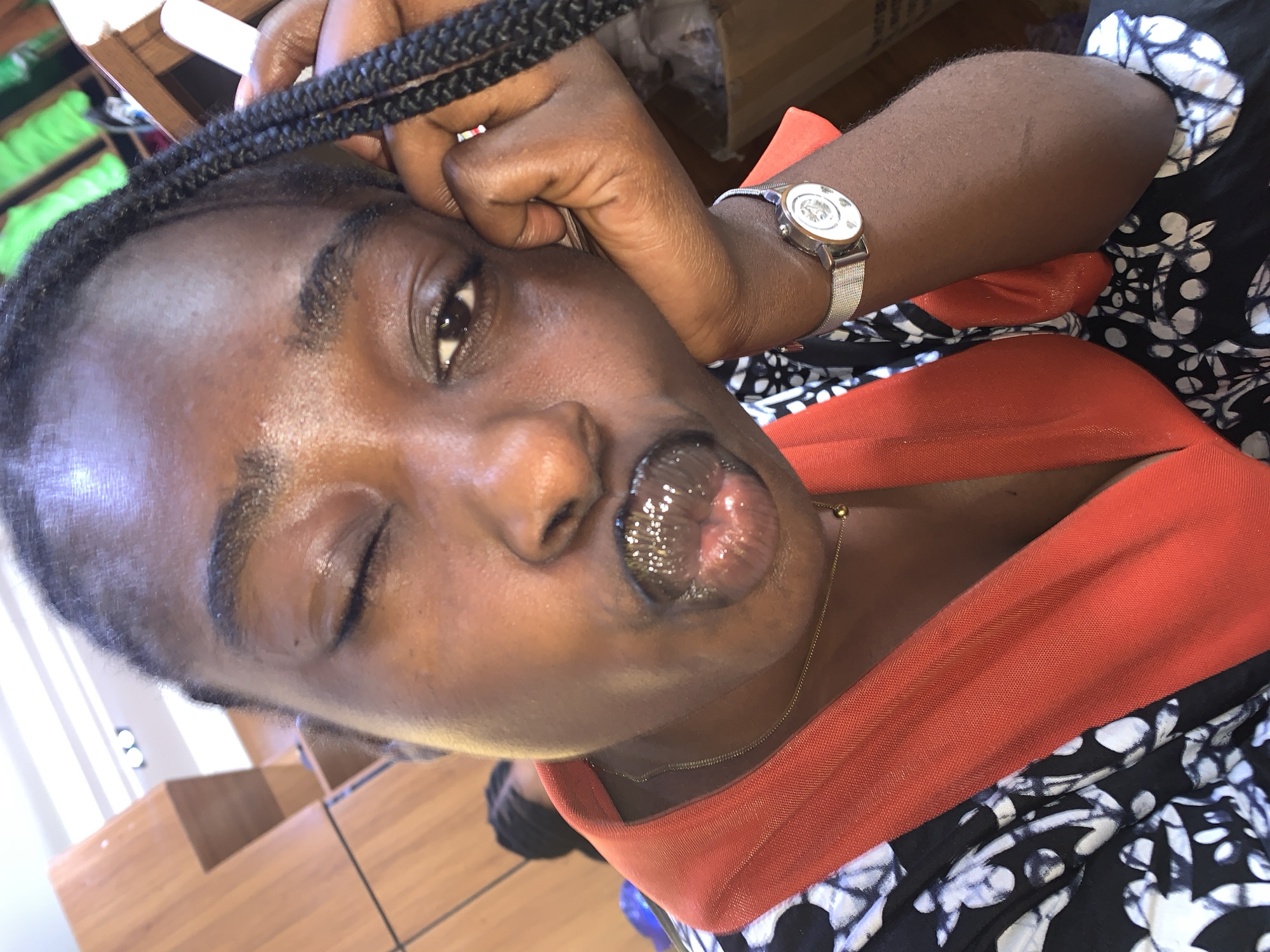 A close-up, head-and-shoulders shot shows a young Black woman winking and making a pout with her lips, her right arm propped on her chin. She has braided hair, and is wearing a black and white patterned garment with an orange neckline. A thin gold chain with a small pendant rests on her collarbone. A silver watch with a white face and silver band is on her left wrist. The background is blurry, showing shelves with what appear to be green items and part of a brown wooden structure. The lighting suggests an indoor environment. The overall impression is a casual selfie taken in a well-lit interior space, possibly a retail or storage area. The specific location of Gura Top, Nigeria cannot be determined from the image.