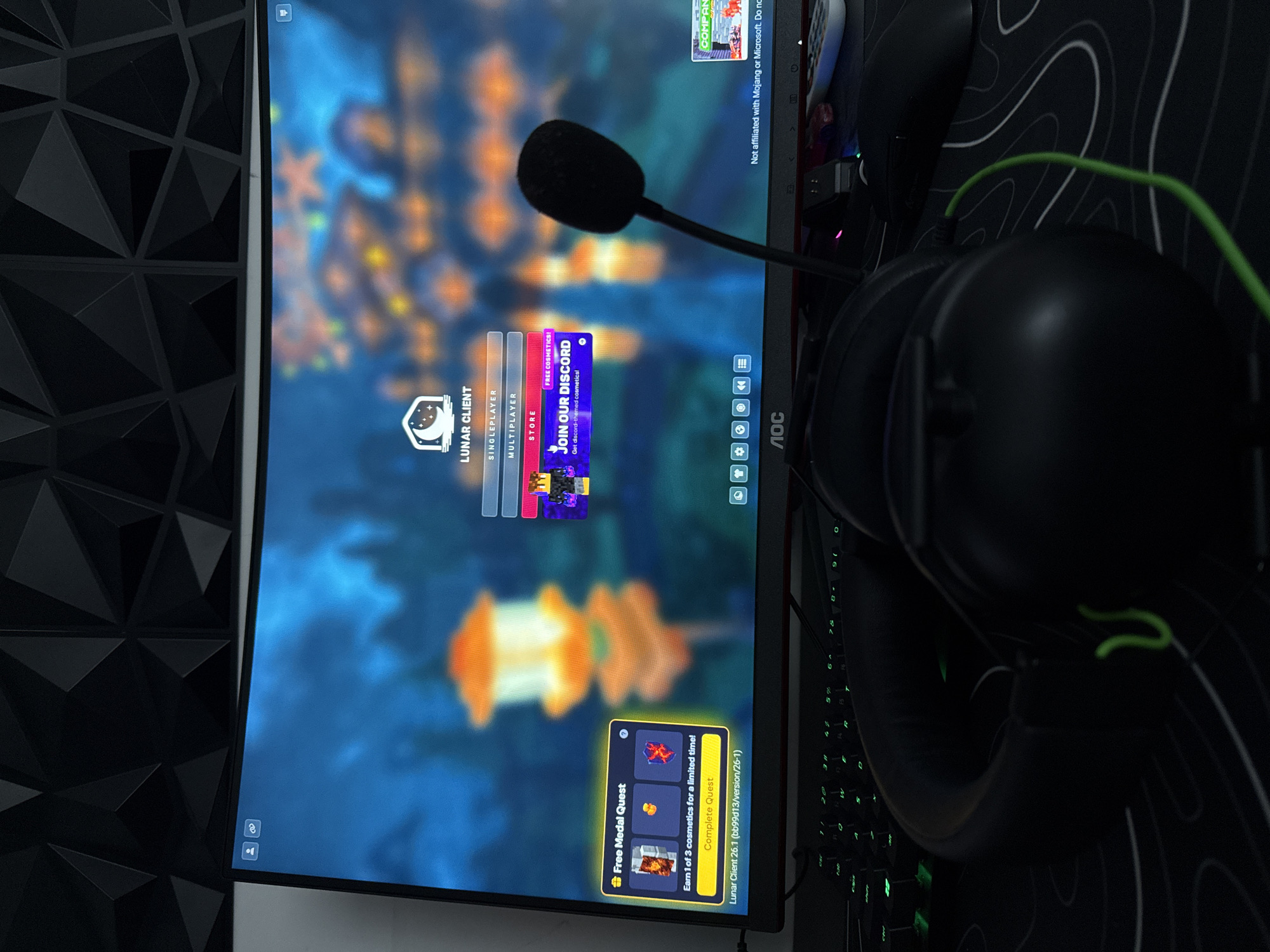 The image depicts a computer setup, likely a gaming station, indoors. A monitor displaying the Lunar Client menu is the focal point. The interface includes options like Singleplayer, Multiplayer, and Store, with a promotional banner for joining their Discord. A black microphone is positioned in front of the screen. To the right of the monitor, a black keyboard with green backlighting is partially visible, along with black headphones. A mouse and mousepad are also present. A textured black wall panel is visible on the left. The environment is dark, with the focus on the illuminated monitor and keyboard. The location is not discernible from the image.