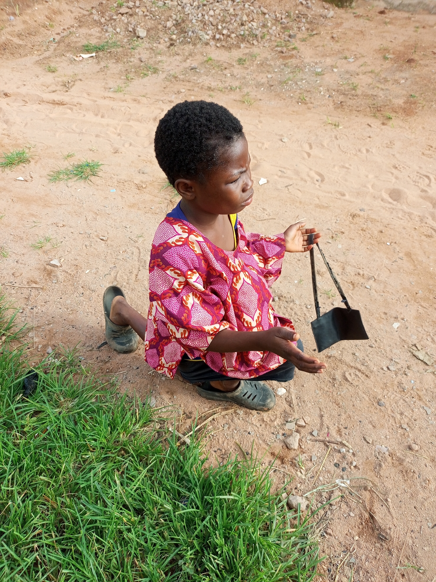A young Black girl is kneeling on sandy ground covered with sparse grass and scattered rocks, in what appears to be an outdoor area within the city of Tempure, Nigeria. The girl is wearing a brightly colored, patterned shirt and dark shorts, with dark shoes on her feet. She is holding a small black object, possibly a wallet or a small bag, in her hands. Her gaze is directed downwards, and her expression is serious. The background consists of more sandy terrain with rocks and some patchy vegetation.