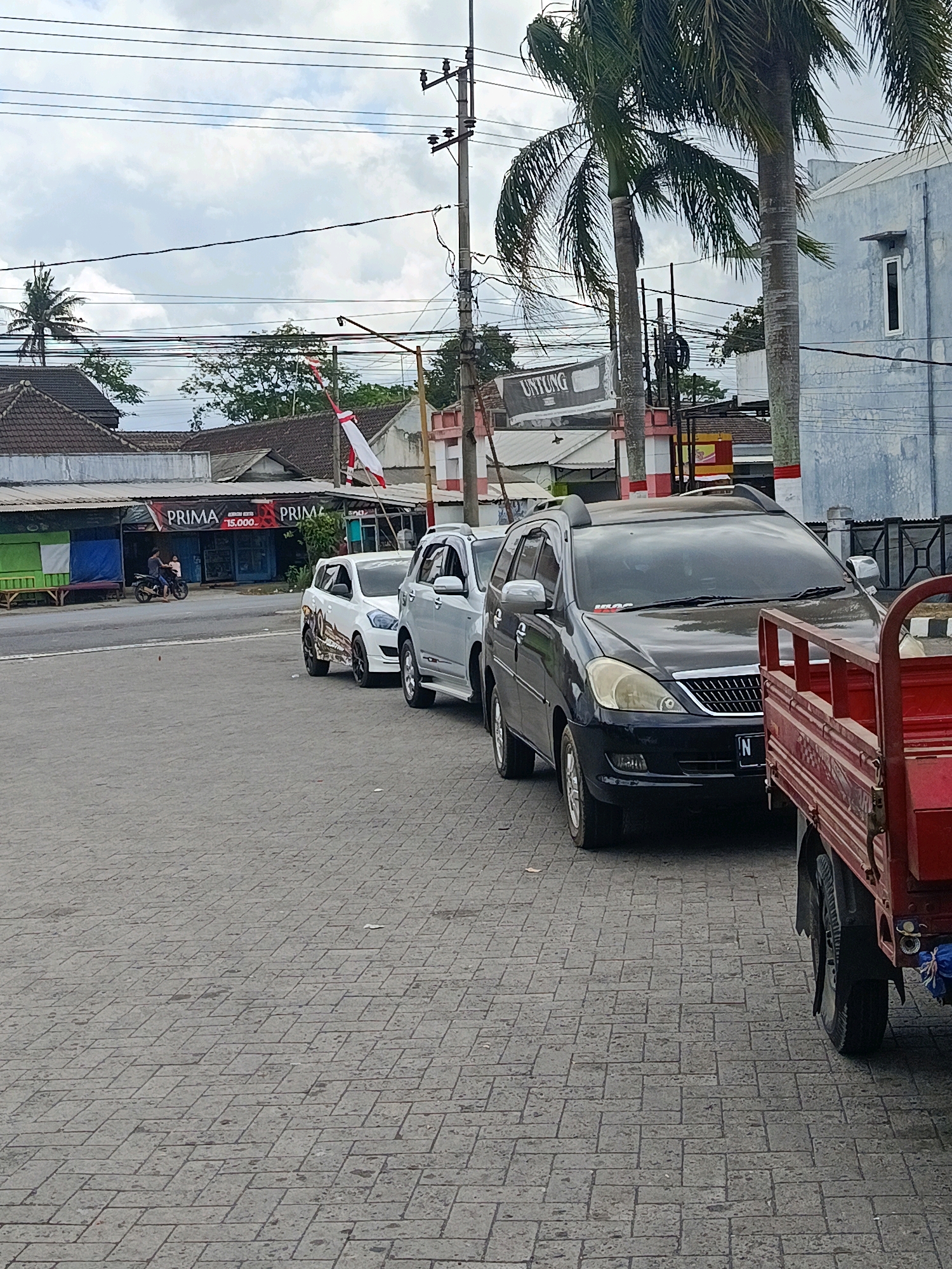 In Tempeh Lor, Indonesia, a street scene unfolds under a cloudy sky. A dark-colored SUV is parked on the right side of the frame, its front end angled towards the viewer. To its left, a white SUV is parked, followed by another white vehicle. A motorcycle with two occupants is visible in the distance, moving away from the viewer. In the background, a row of buildings lines the street, some with awnings and signage. A red truck is partially visible in the foreground on the right. The ground is paved with interlocking stones. Overhead, power lines crisscross the sky, supported by utility poles. Palm trees are scattered throughout the scene, some reaching high into the sky.