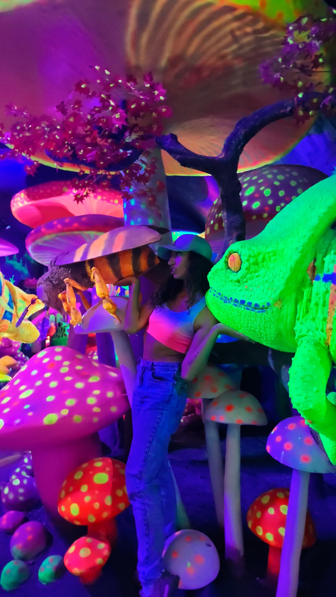 A woman stands among large, brightly colored, luminescent mushroom decorations and a giant lizard in what appears to be an indoor, artificial mushroom forest setting. The woman is wearing a tie-dye crop top, jeans, and a baseball cap. She is standing between an oversized, painted bee on a mushroom and a large, green, cartoonish lizard with orange eyes. The scene is heavily illuminated with ultraviolet light, which causes the neon colors of the decorations to glow intensely. The location is possibly a commercial attraction with a whimsical, fantasy theme in the city of Binh Lu, Vietnam.