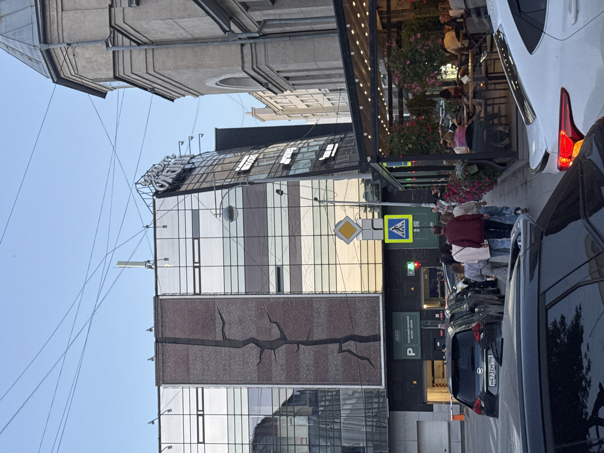 The image depicts a street scene in Yekaterinburg, Russia. In the foreground, several cars are parked or in motion on a paved road. A black SUV with the license plate "M473EM 196" is prominently visible. To the right, a white car's taillight is illuminated. The middle ground features a modern building with a glass facade and a large, dark vertical design resembling a crack. Beneath this building, there is an entrance to a parking garage with a sign indicating "ПЛАТНЫЙ ПАРКИНГ 07:00-23:00" (Paid Parking 07:00-23:00). People are walking along the sidewalk. To the right of the modern building, an older, stone building with arched windows stands. Adjacent to the modern building's ground floor is an outdoor seating area for a cafe or restaurant, adorned with hanging plants and lights, with patrons seated at tables. A pedestrian crossing sign and a yield sign are visible on the street. The sky is clear and blue.