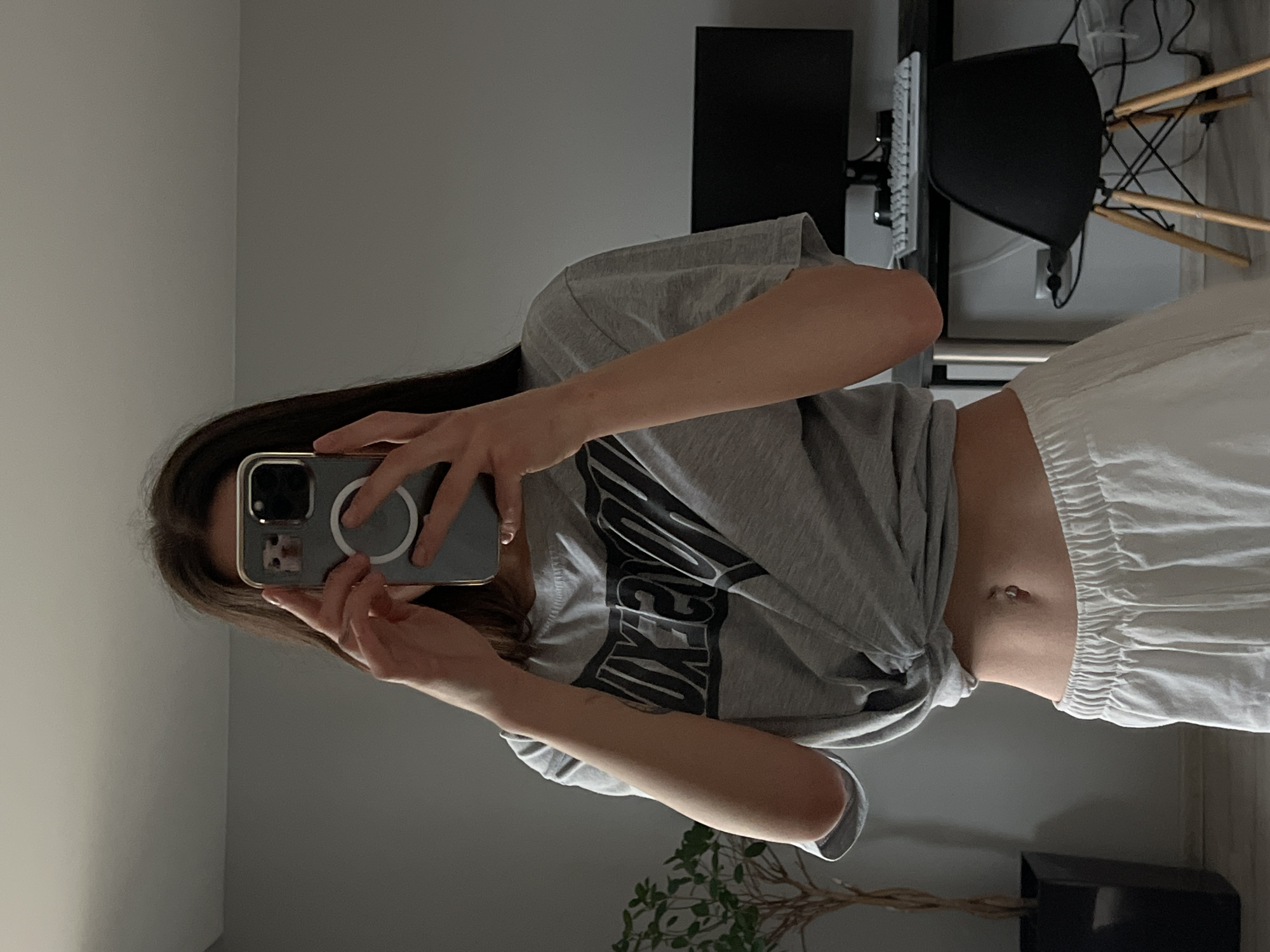 A young woman is captured in a full-length mirror selfie, her face obscured by the phone she holds. She has long, dark hair and is dressed casually in a grey t-shirt with the word "NIKE" emblazoned in large black letters across the front. The t-shirt is tied at the waist, revealing her midriff, which features a navel piercing. She is also wearing loose-fitting white pants. Her phone, likely an iPhone, is encased in a grey protective cover with a visible white MagSafe ring.
The setting appears to be an indoor room, possibly a bedroom or home office. In the background, a dark computer monitor or television screen is visible on a black desk, alongside a white keyboard and numerous wires. A black office chair with wooden legs is positioned near the desk. The room features light grey walls and a dark rectangular pot containing a plant with bare branches in the foreground. The lighting is soft and ambient, indicating an indoor environment, without direct sunlight. The scene suggests a modern, functional living space in Ternopil, Ukraine.