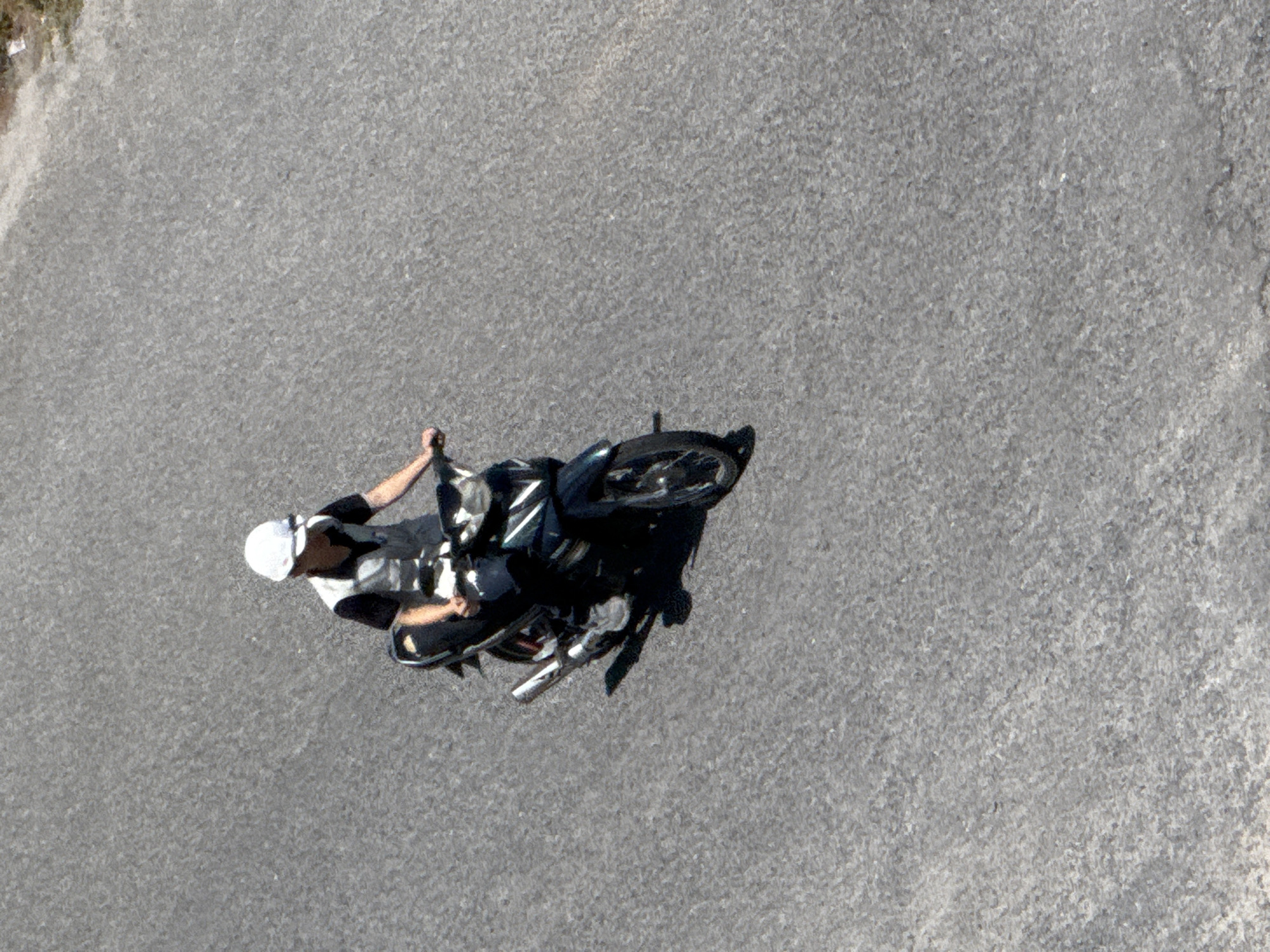 An aerial shot shows a person on a motorcycle, viewed from directly above. The person is wearing a white helmet and dark clothing, with a white and black striped shirt. They are seated on a black motorcycle, which is angled to the right in the frame. The motorcycle has visible chrome accents and black tires. The scene is set on an asphalt surface, which appears to be a road or a paved area. The texture of the asphalt is rough and grey, with some lighter patches. There are no other people or vehicles visible in the immediate vicinity. The lighting suggests it is daytime, with bright sunlight casting shadows, though the overall weather is not explicitly discernible. The location is identified as Nha Trang, Vietnam, but there are no specific visual cues in the image to confirm this. No text is visible in the image. The activity appears to be the person riding or stopped on the motorcycle. The perspective gives a sense of openness, with the vast grey expanse of the asphalt dominating the background.