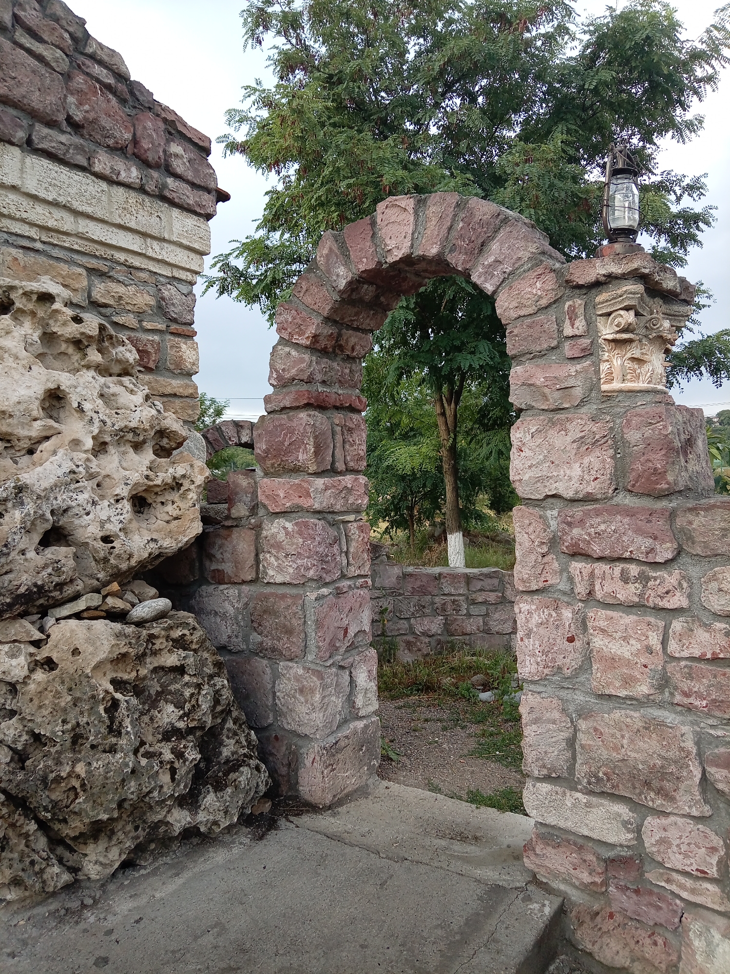 This is a photograph of an ancient stone archway, likely a remnant of a historical structure. The arch is constructed from rough-hewn reddish-pink stones, giving it a rustic appearance. To the left of the arch, a large, irregularly shaped boulder with visible pores and crevices is stacked against a wall made of similar stone, with some lighter-colored, smoother stones layered horizontally. To the right of the arch, the stone wall continues, featuring decorative carvings and a hanging lantern with a glass enclosure at the top. Behind the arch, a lush green tree with dense foliage dominates the background, its branches reaching towards a pale, overcast sky. The ground in front of the arch appears to be paved with concrete, showing some wear and tear. The overall scene suggests an outdoor historical site or ruin, possibly in a park or garden setting. The presence of the lantern and the decorative carvings implies a deliberate artistic or architectural element, rather than a purely utilitarian structure. The image captures a sense of age and enduring history through its weathered materials and time-worn architecture. The context provided places this scene in Yessentuki, Russia.