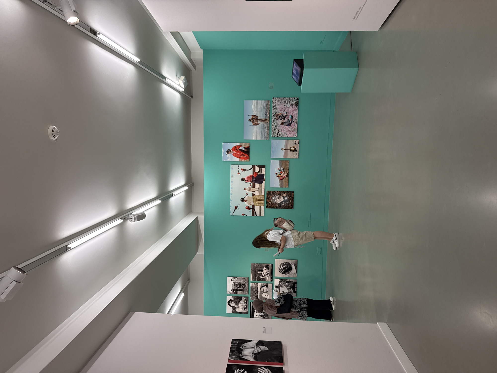 This image depicts an indoor art exhibition, likely a photography exhibition given the displayed works. The walls are painted in a vibrant teal color, contrasting with the white walls on the left and right. The floor is a polished grey concrete. Several people are present, appearing to view the artworks.
On the left wall, two framed black and white photographs are partially visible, showing close-ups of hands and a person's face. In the foreground on the left, a person wearing a hijab and a patterned top stands facing the teal wall, seemingly engaged with the artwork.
The teal wall displays a grid of various photographs. These appear to be color prints and are of different sizes. The subject matter of the photographs seems diverse, including portraits of people in traditional attire, scenes of people by water, and close-ups of individuals.
In the center right, another person, dressed in a white t-shirt and tan shorts, is standing and looking at a piece of paper, likely an exhibition guide or artwork description, while facing the teal wall.
Further to the right, a light blue pedestal supports a dark electronic device, possibly an interactive display or audio guide. The ceiling features a track lighting system with multiple spotlights and fluorescent tubes, providing illumination for the exhibition space.
While the specific location within Frankfurt, Germany, cannot be determined from the image alone, the context suggests a public art venue such as a gallery or museum. The overall impression is that of a modern and well-lit exhibition space designed for patrons to appreciate visual art.