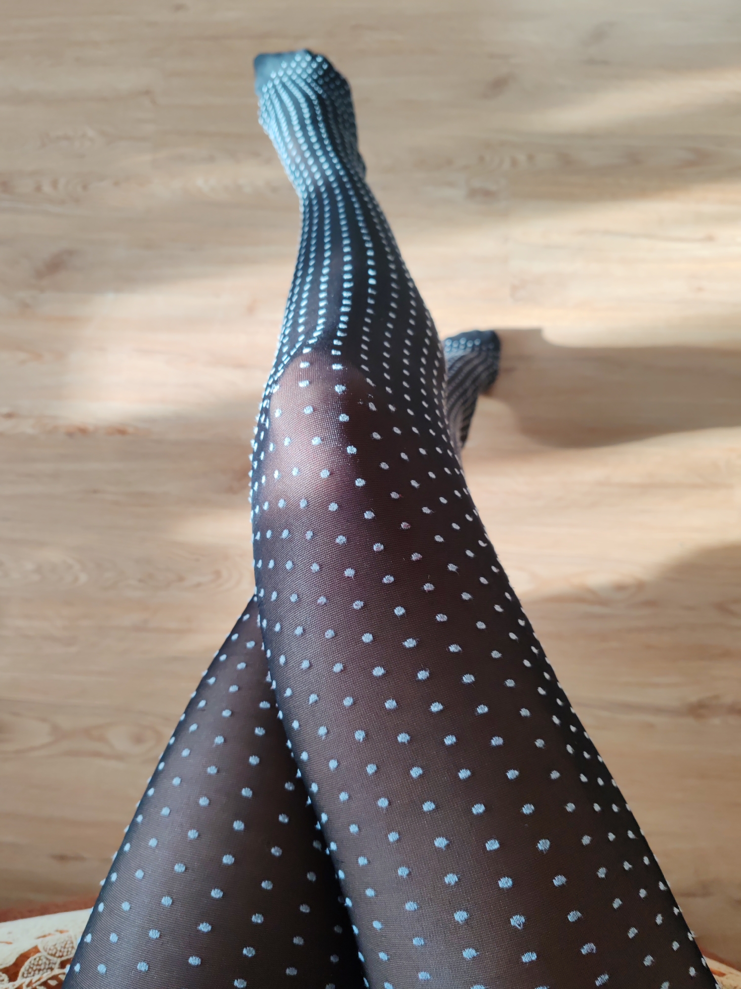 The image shows a pair of legs crossed at the ankles, wearing black sheer tights with small white polka dots. The legs are positioned on a light brown wooden surface. The background is blurred, but appears to be a room interior. There is no information to indicate the location is Petropavlovsk-Kamchatsky, Russia. No people besides the legs are visible. The primary object is the tights-clad legs. The action is the crossing of the legs. The environment is an indoor space.