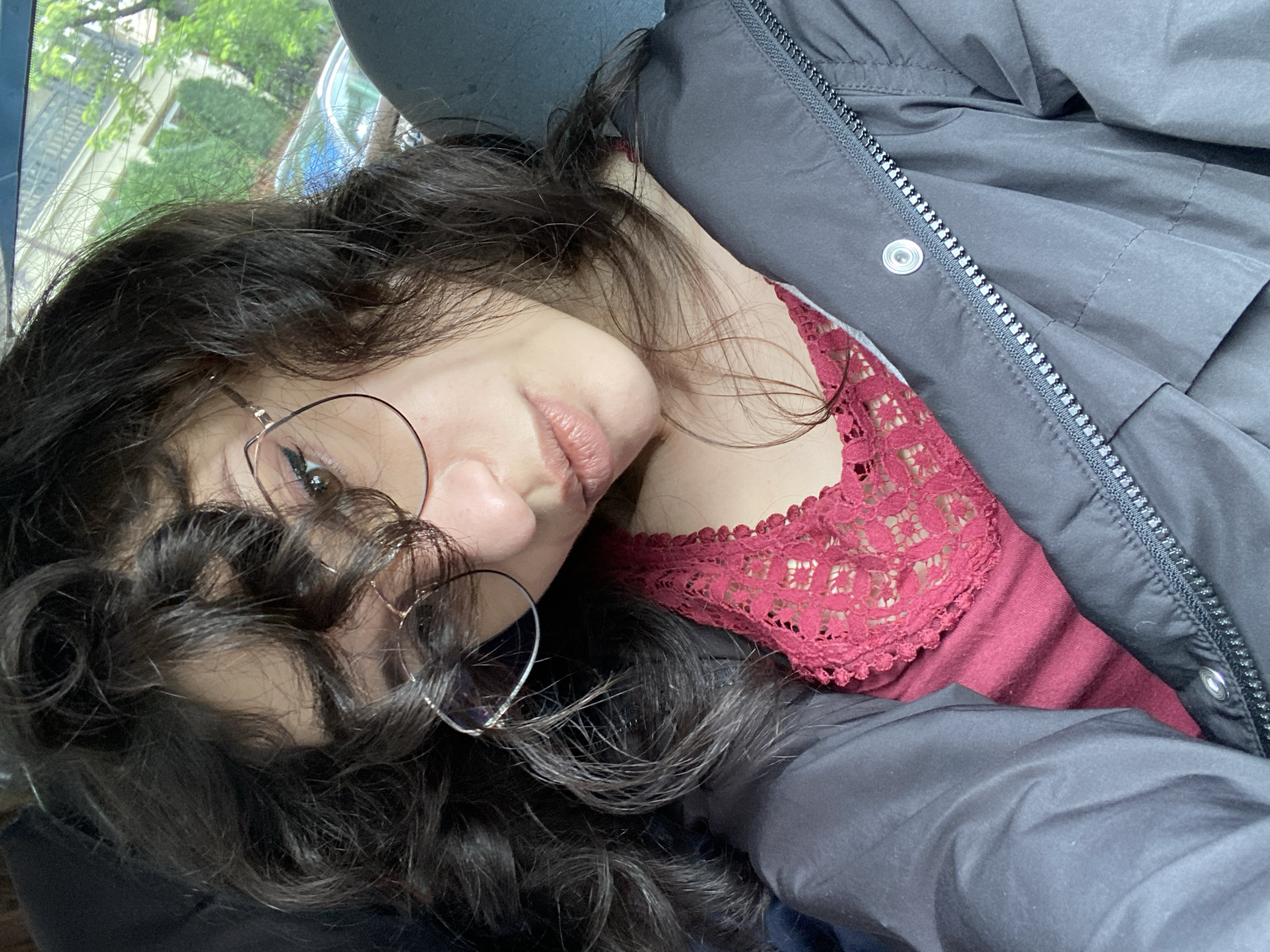 A close-up, selfie-style photograph features a young woman with dark, curly hair and glasses. She is wearing a dark jacket over a dark red top with lace detailing. The background appears to be a car interior, with a glimpse of a street scene outside the window, including buildings and trees, suggesting an urban environment. The overall lighting suggests it is daytime. There is no discernible evidence to locate this image in Sofia, Bulgaria.