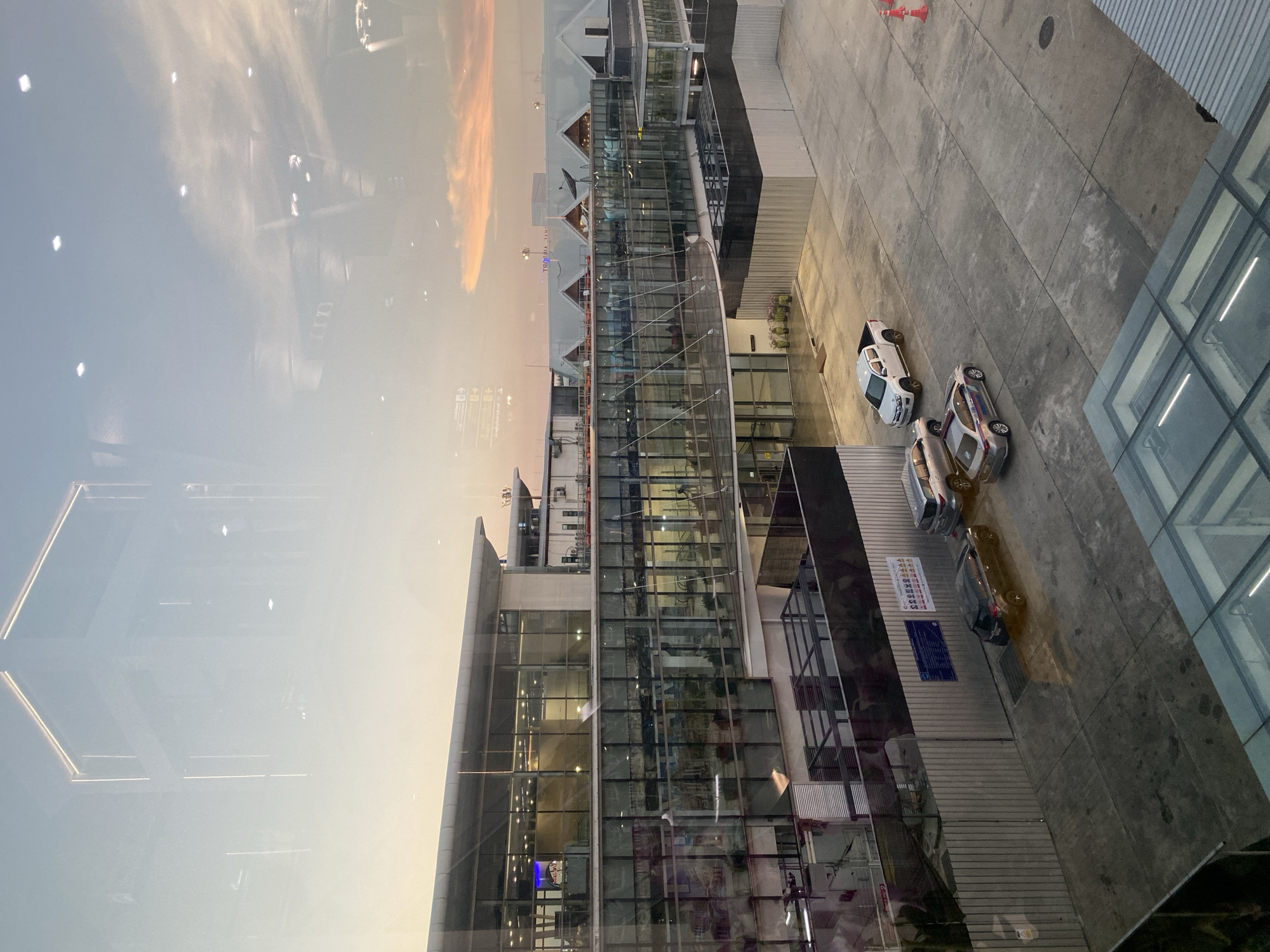 The image depicts an elevated perspective of a modern, multi-story building with extensive glass facades, potentially a hotel or commercial complex, located in Mai Khao, Thailand. The architecture includes distinct gabled roof sections and connecting glass walkways. The scene is viewed through a window, with the sky visible in the reflection. The sky shows a mix of blue, white, and orange hues, indicating either sunrise or sunset. Adjacent to the building is a paved area with three parked vehicles, and what appears to be an airport entrance for pick up and drop off. A sign and safety cones are also visible on the pavement. The overall impression is of a contemporary architectural design integrated into the urban landscape of Mai Khao, Thailand.