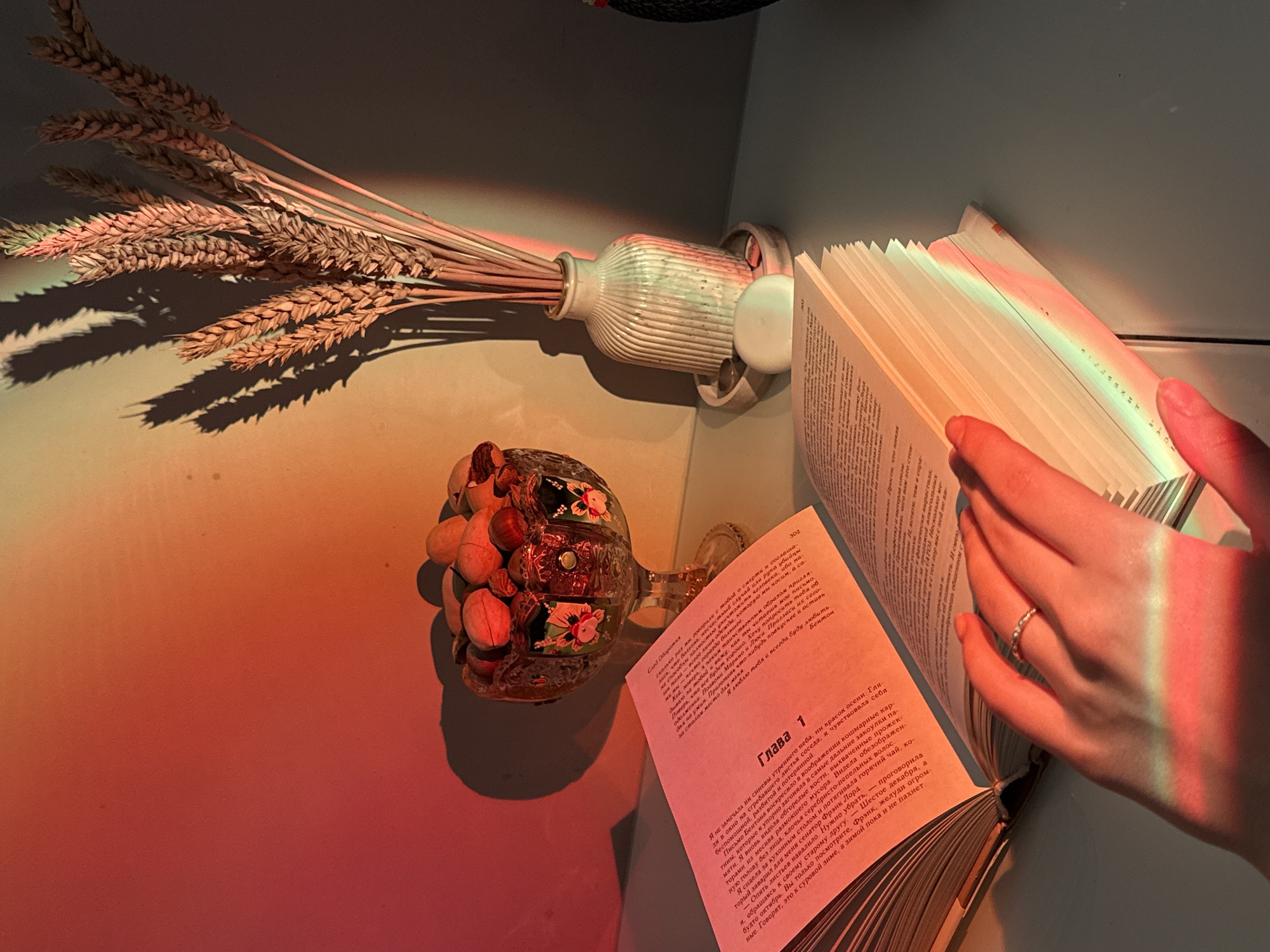 The image captures a cozy indoor still life with a strong, atmospheric play of light. A person's hand, adorned with a silver-colored ring, is holding an open book, suggesting an act of reading. The scene is split by dramatic lighting: one side, featuring a bouquet of wheat stalks in a white ribbed vase and a decorative glass goblet filled with nuts or small items, is bathed in a warm, reddish-orange glow, casting long, defined shadows. The other side, where the book and hand are, has cooler, blue-toned ambient light, overlaid with a vibrant rainbow-colored stripe that extends across the book's pages and the reader's hand, indicating light refraction.
The book is open to a page with the heading "Глава 1" (Chapter 1) in Russian. Visible text on the left page includes "След Оборотня" (Trail of the Werewolf) and mentions "Бентон" (Benton). The right page details a narrative about feeling "helpless, broken, and lost," referencing "Benton's letter," "nightmarish pictures," "burnt bones," and a character named "Senator Frank Lord." It also mentions "December sixth" and thoughts about winter. The overall setting appears to be a personal space, possibly a desk or a bedside table, accentuated by the decorative elements and the intimate act of reading. The distinct lighting creates a dreamy and focused ambiance.
