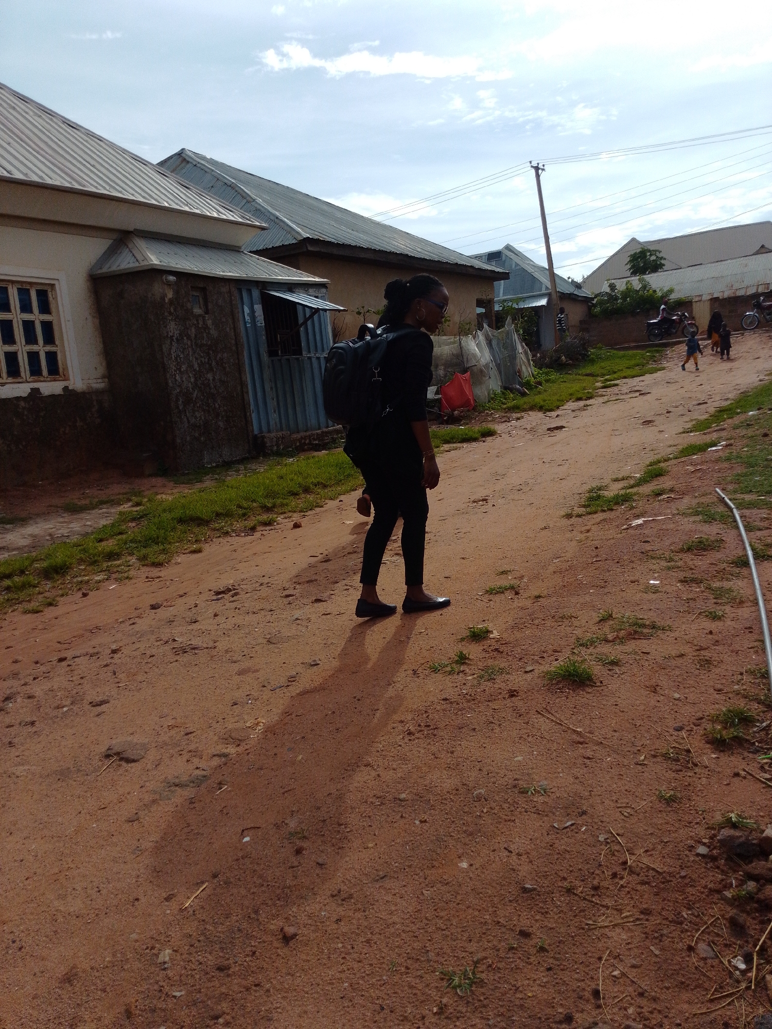In Gombe, Nigeria, a young Black woman with dark hair pulled back and wearing glasses walks down a dirt road, her back to the camera. She is dressed in black pants and a black shirt and carries a black backpack. Her shadow stretches out on the reddish-brown dirt road, which is uneven with scattered stones and patches of dry grass. To her left, several buildings with corrugated metal roofs are visible. One building has a light-colored exterior with a multi-paned window. Adjacent to it is a structure with a textured gray wall and a metal-covered entrance. Further down the road, other buildings are seen, along with utility poles and wires. In the distance, a few people and motorcycles are visible, suggesting ongoing activity. The sky is a light blue with scattered white clouds. The overall scene depicts a residential area in Gombe, Nigeria, on a partly cloudy day.