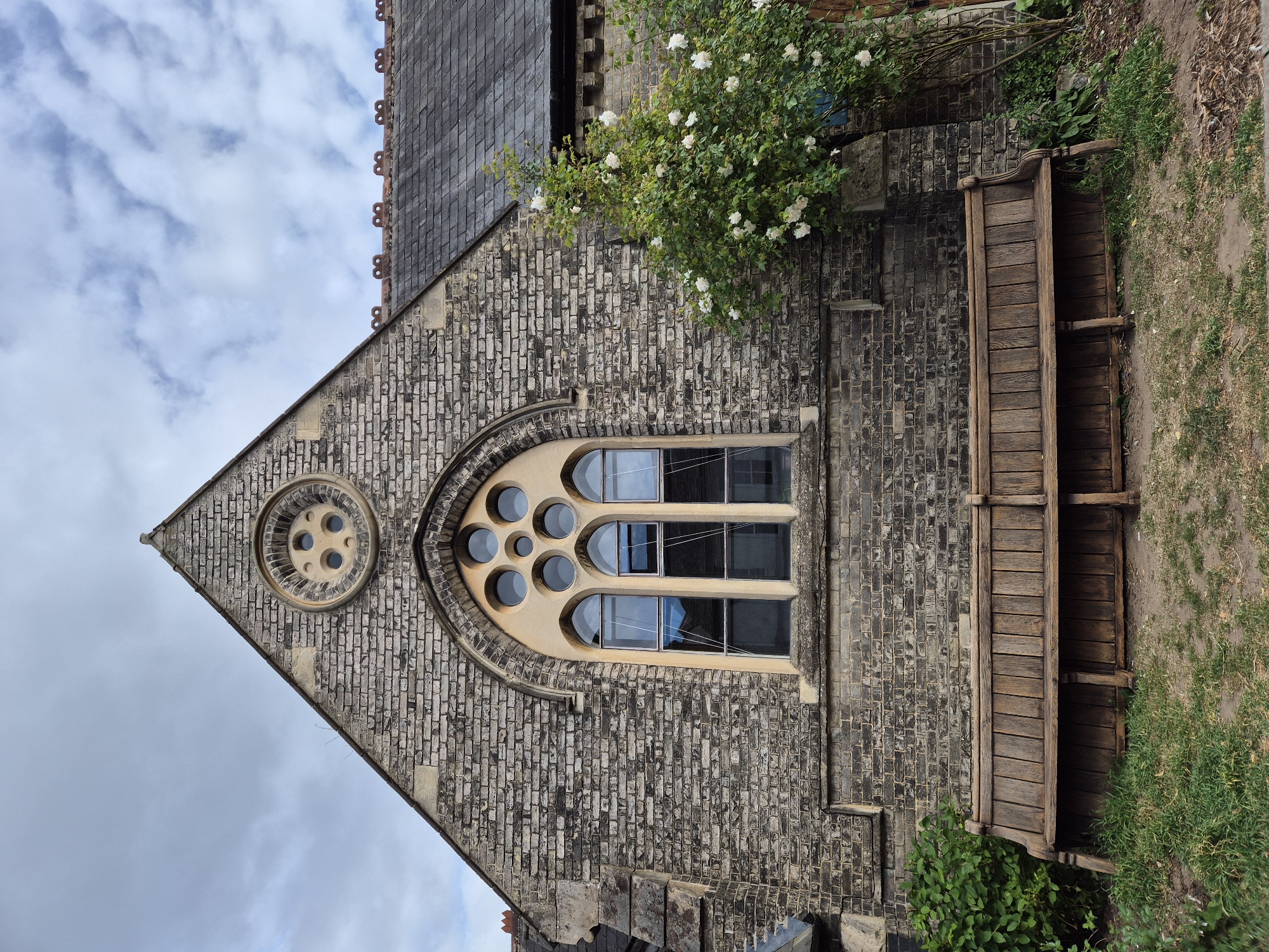 The image shows the exterior of a brick building, likely a church, in West Suffolk, United Kingdom. The building features a steeply pitched roof with slate tiles. A triangular section of the facade has a circular window with multiple smaller round windows. Below this, a large arched window with rectangular panes is visible. A wooden bench sits against the wall adjacent to the window. A flowering bush grows on the wall above the bench. The sky is visible in the background, with patchy cloud cover.