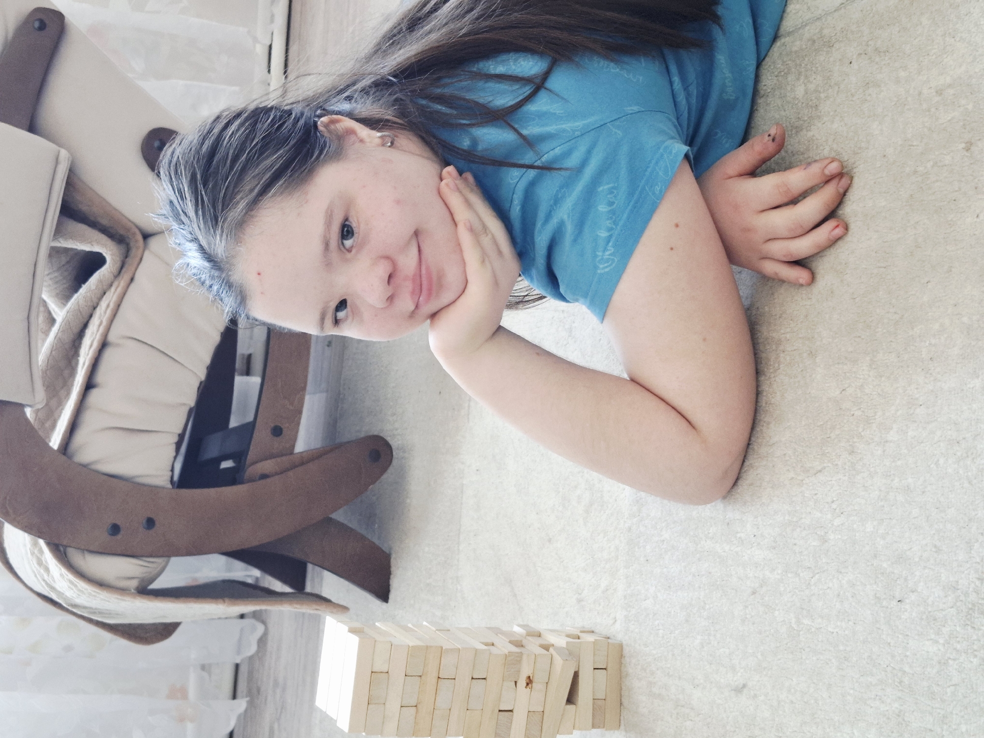 A young woman is lying on the floor next to a wooden chair and a stack of wooden blocks. The woman is wearing a blue shirt and resting her head on her hand, smiling at the camera. The wooden blocks are arranged in a tower. The location is likely Chaikovsky, Russia, based on the available information.