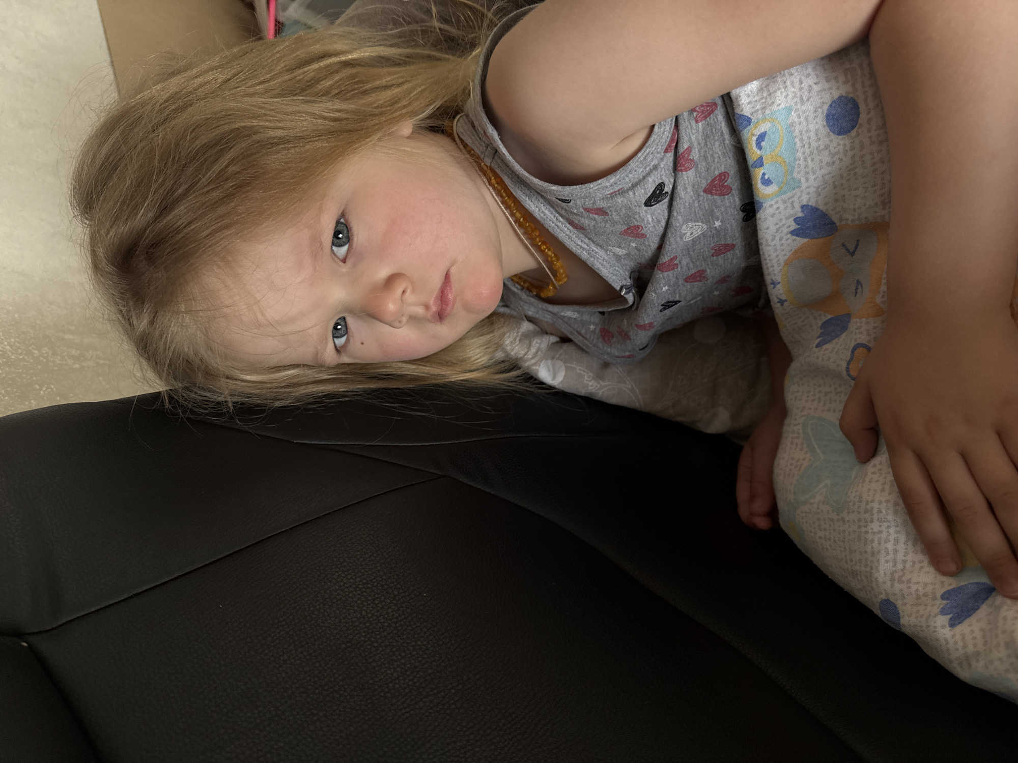 A young girl with long, light blonde hair and striking blue eyes is the central subject of the image. She is lying on her side, resting her head on a dark, textured surface, likely a car seat or a couch. Her gaze is directed straight at the camera, with a neutral to slightly pensive expression; her cheeks show a light flush.
She is dressed in a gray t-shirt adorned with outlines of red, black, and white hearts. Around her neck, she wears a light amber bead necklace. Part of her body, extending from her waist downwards, is covered by light-colored pajamas or blankets featuring an owl pattern with blue and orange accents. Her hands are visible resting on the patterned fabric.
The background is indistinct, showing a lighter, possibly off-white, surface. The lighting suggests an indoor setting. There are no clear indicators of the time of day or external weather. No visible text is present in the image. The scene is located in an indoor setting, potentially in Ферма, Russia.