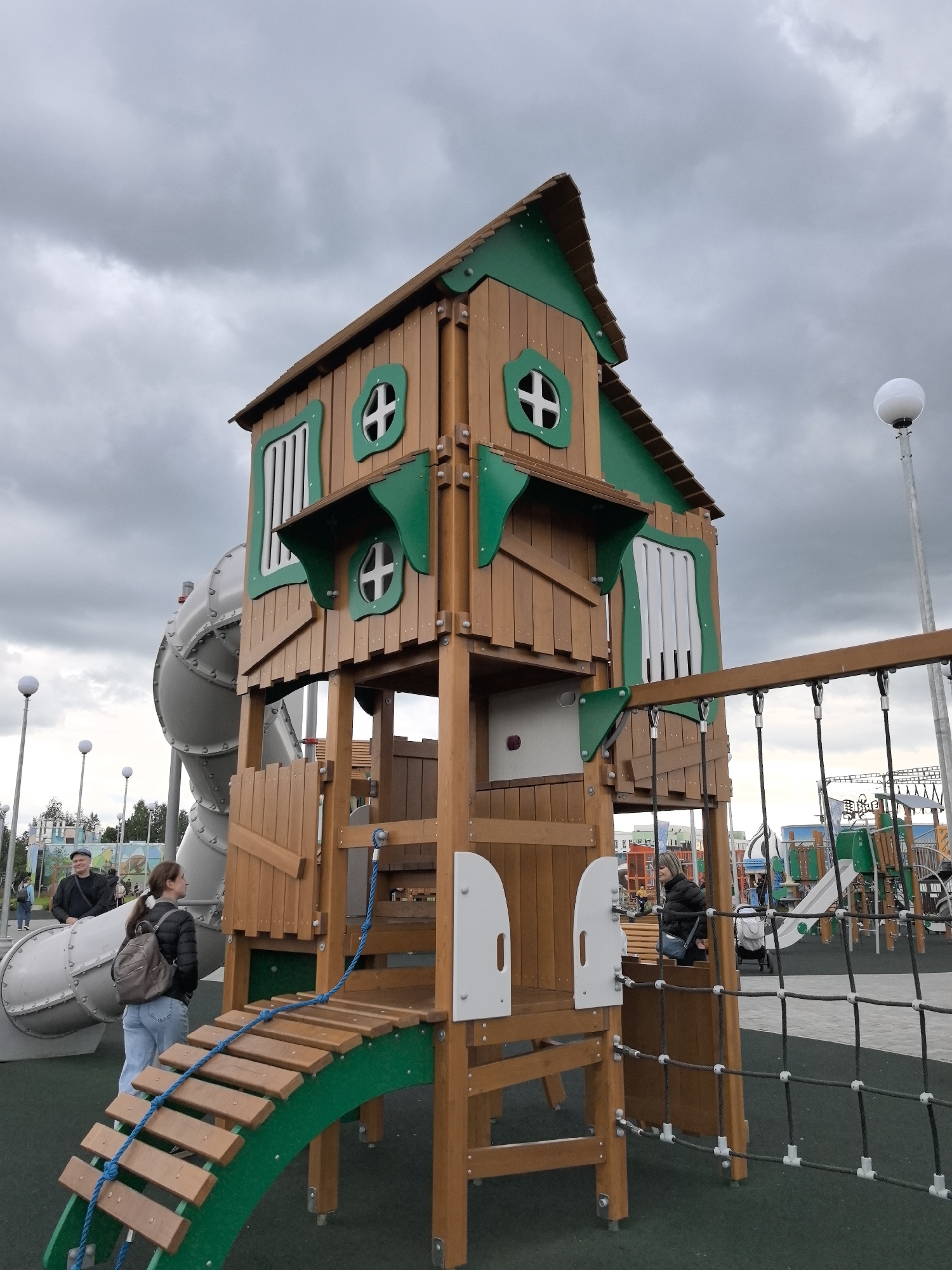 The image shows a playground structure resembling a wooden treehouse in the city of Perm, Russia. The structure is two stories tall, with a green roof and green trim around the windows and doors. The windows are circular with a cross design. A woman with a backpack stands near the base of the treehouse, next to a large silver slide. A man in a cap stands near the slide as well. A net climbing structure is attached to the side of the treehouse. In the background, there are additional playground equipment and other people. The sky is overcast with gray clouds. Lamp posts are visible in the background.