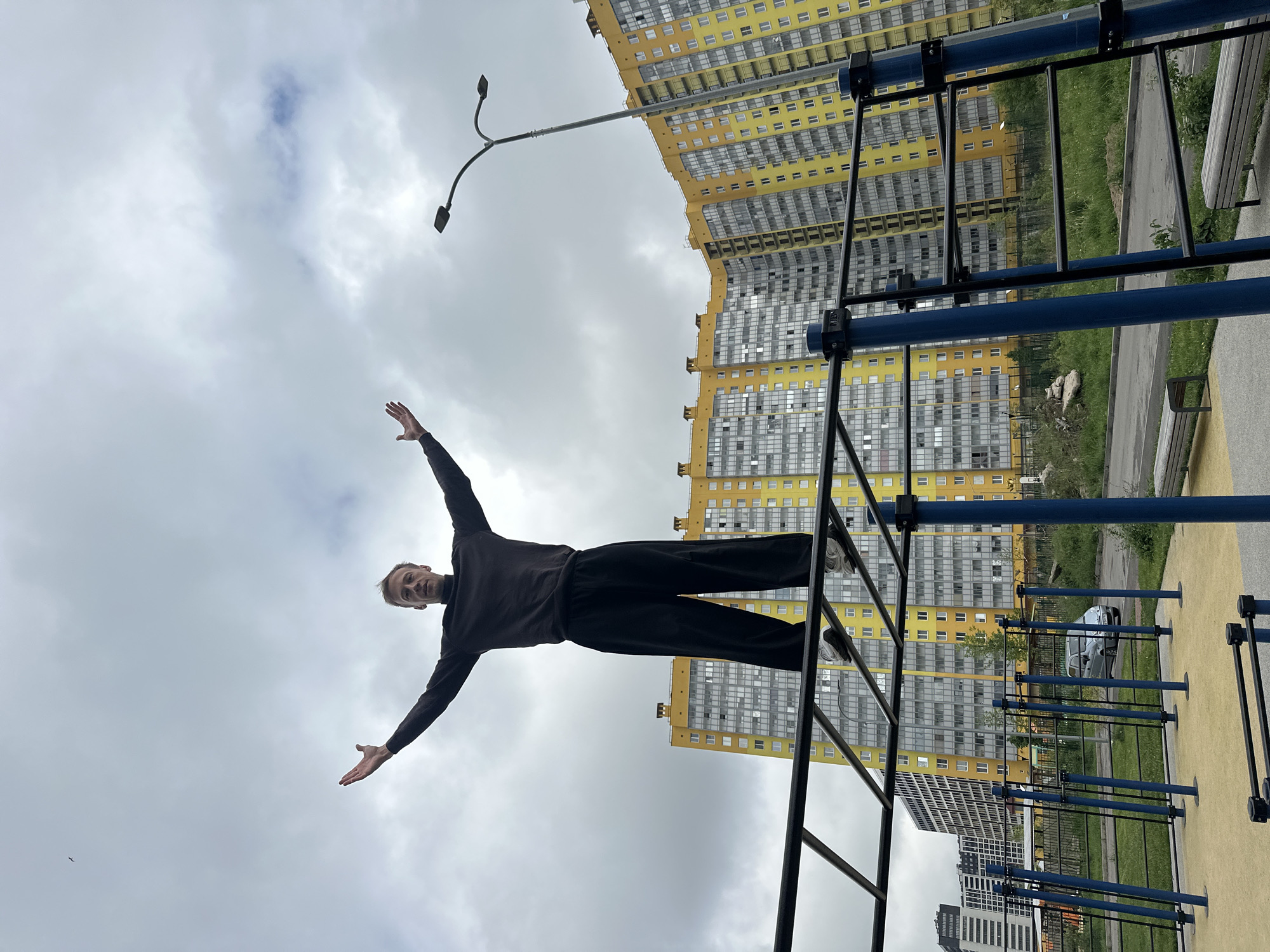 A man stands atop a blue and black metal climbing structure in Murino, Russia. He wears dark pants and a dark long-sleeved shirt, and his arms are outstretched. The background includes tall, yellow and gray apartment buildings under a cloudy sky. A streetlight is visible. Ground level amenities such as a bench and a fence. The image appears to have been rotated to a vertical view.