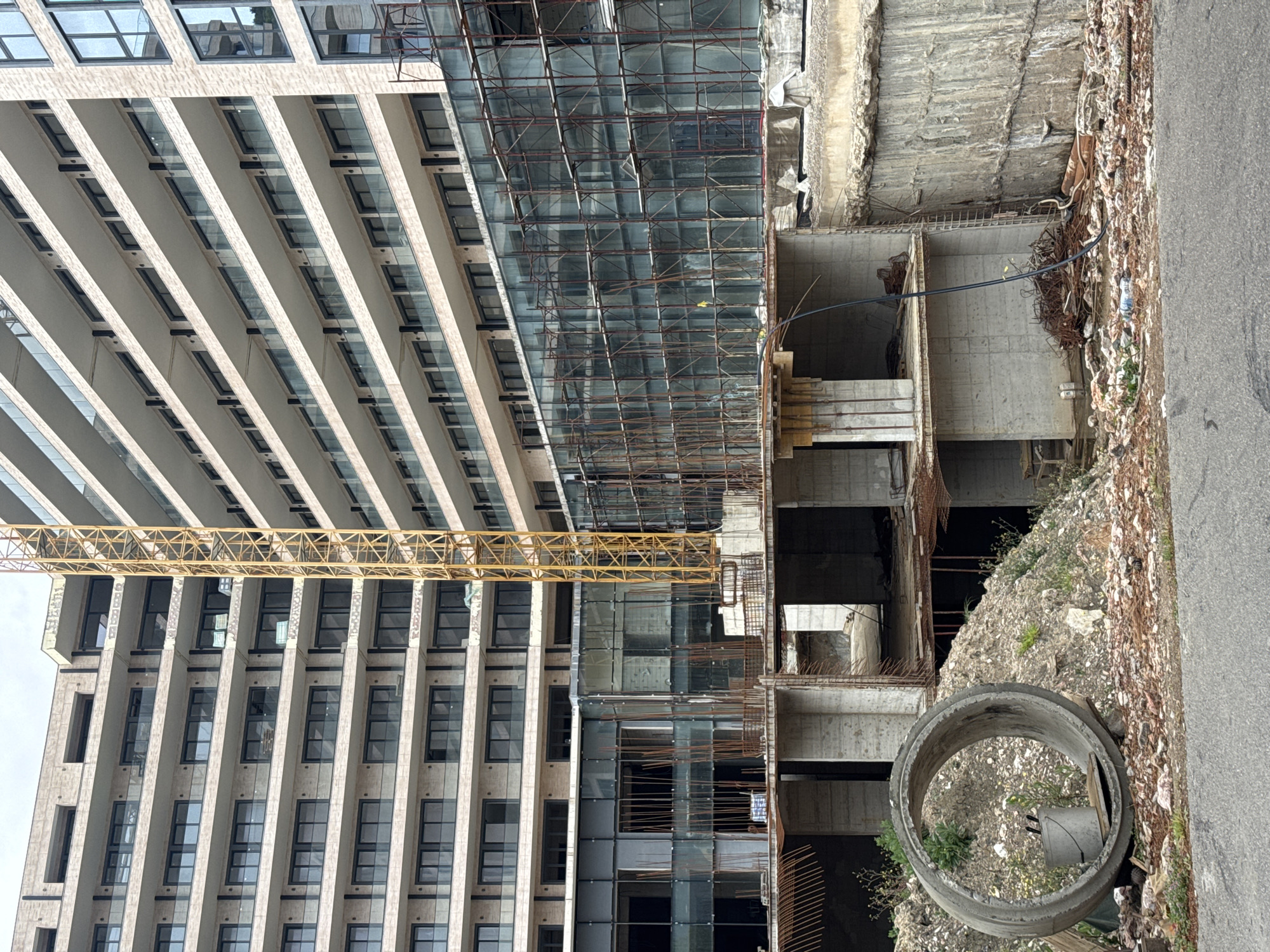 The image depicts a construction site in Budva, Montenegro. A high-rise building with multiple stories is partially constructed. A yellow crane arm extends across the building's facade. The construction site shows exposed concrete structures and rebar. A large concrete pipe lies on the ground near the edge of the site. The ground is littered with construction debris and trash. The image suggests an urban environment in Budva.