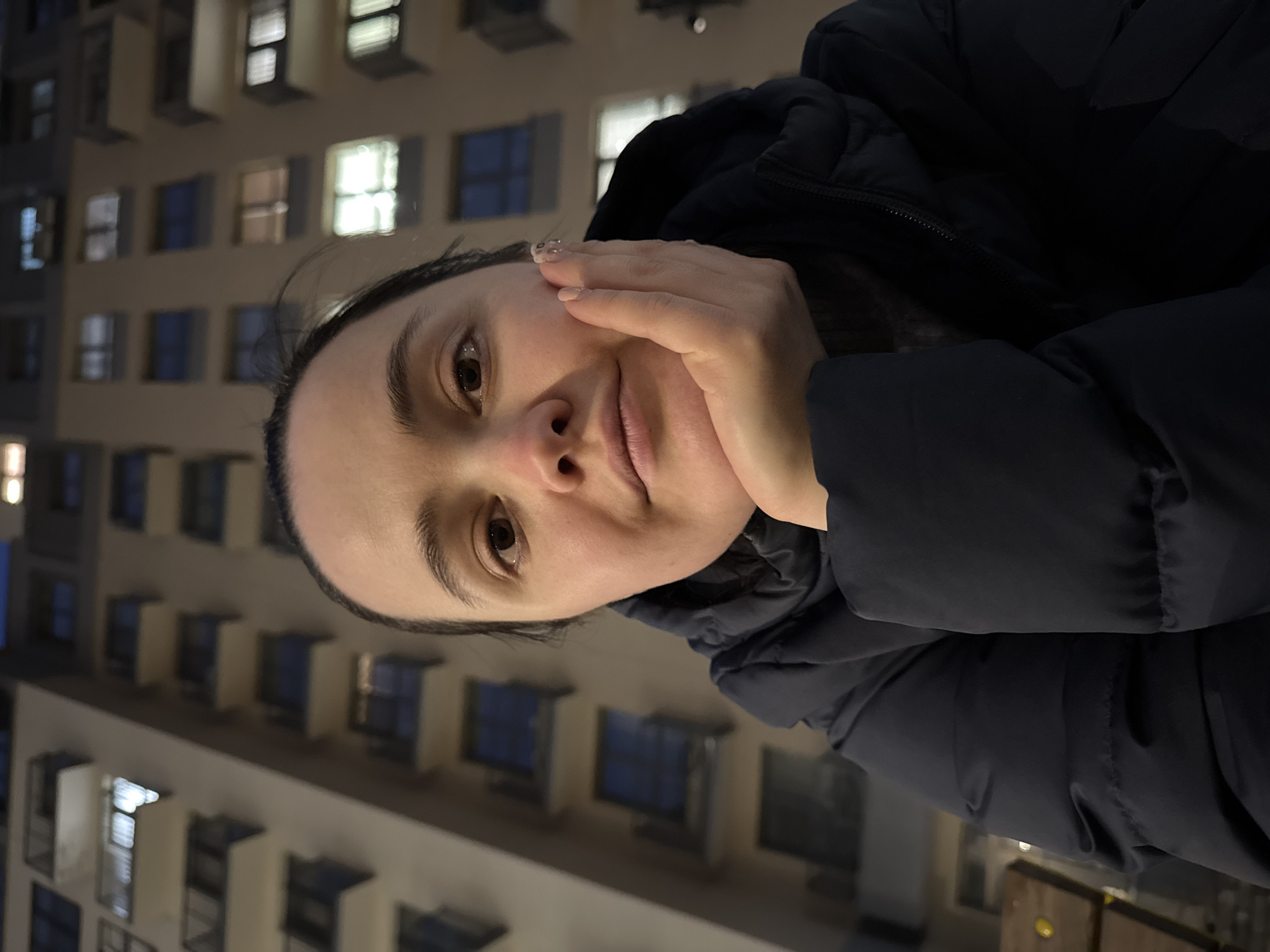 A woman wearing a black puffy jacket is positioned with a light tan building in the background. The building has multiple windows, some illuminated, arranged in a grid-like pattern. The woman is holding her hand to her face. The image appears to be taken at night. While the image exhibits characteristics consistent with urban architecture found in many cities, there's no definitive evidence within the picture to confirm its location as Izhevsk, Russia, or any other specific city.