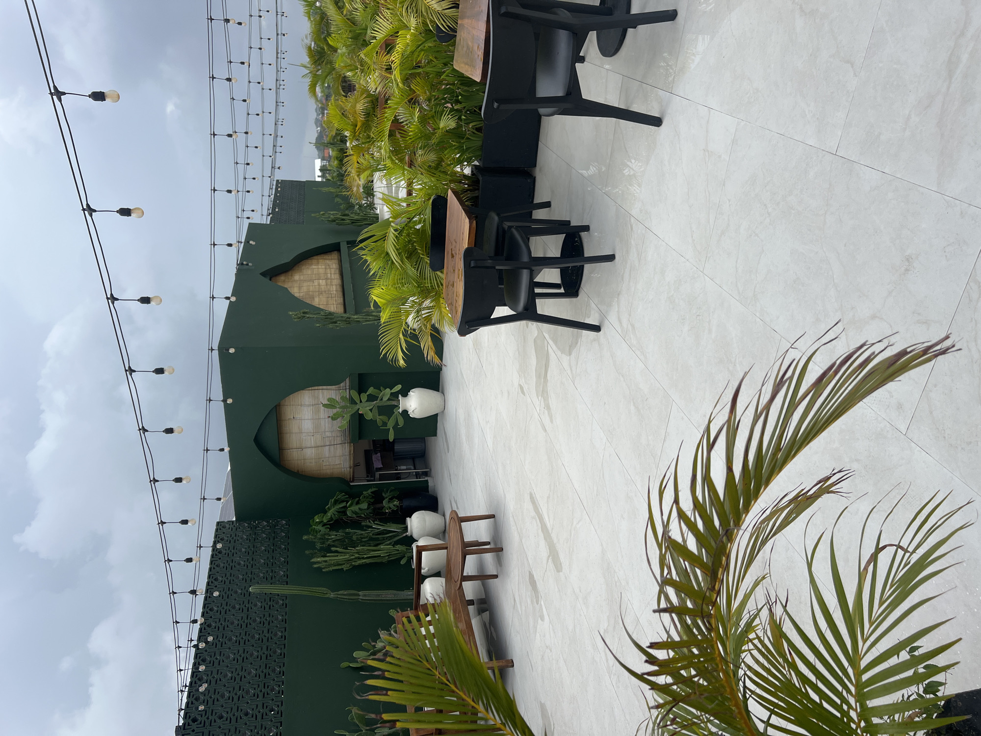 This image depicts an outdoor patio or terrace, likely part of a restaurant or cafe, set in North Kuta, Indonesia. The scene features modern, dark green architectural structures, some with arched openings covered by woven bamboo blinds. A section of one wall displays a dark, intricately patterned decorative screen.
The floor is covered in large, light-colored rectangular tiles that appear damp or wet, reflecting the overcast, cloudy sky above. The space is furnished with several dark, modern dining tables with wooden tops and black chairs. String lights with exposed bulbs are strung overhead, designed to illuminate the area.
Lush tropical vegetation is abundant, including numerous palm plants with vibrant green fronds, various cacti, and other leafy plants. These are arranged in planters and elegant white urns of differing sizes. A wooden bench or low table is also visible amidst the greenery. No people are present, and there is no discernible text. The atmosphere is serene and stylish, indicating a comfortable outdoor dining or relaxation spot under a cloudy daytime sky.