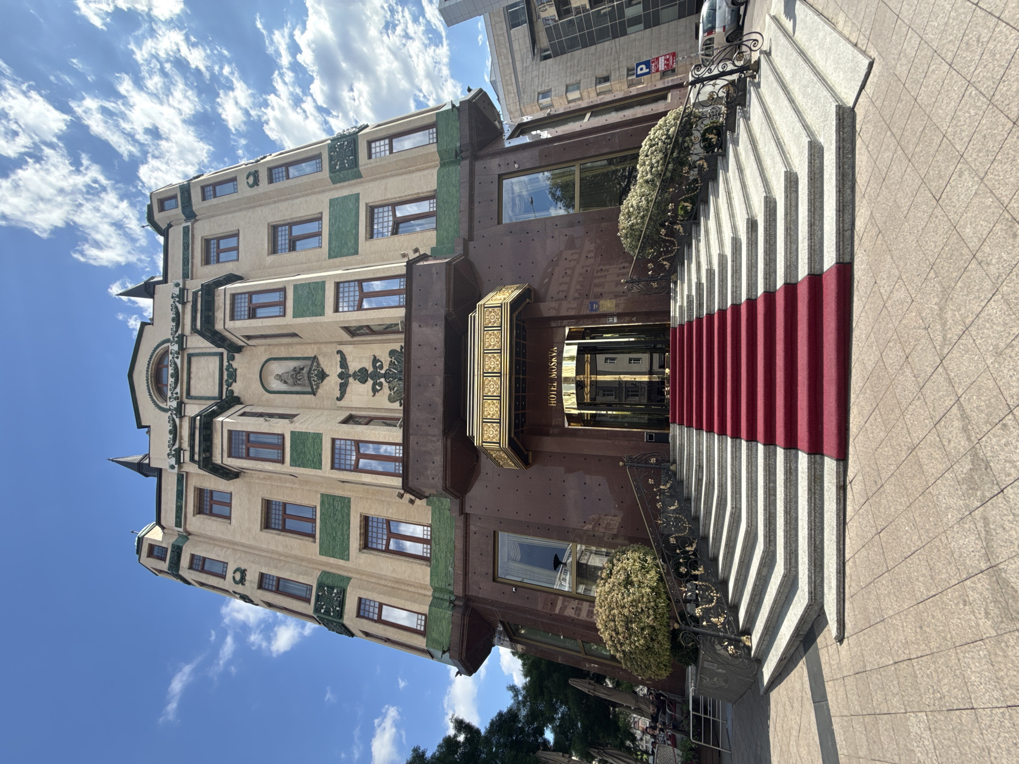 The image shows Hotel Moskva in Belgrade, Serbia. The hotel is a multi-story building with a distinctive architectural style, combining elements of Secession and Art Nouveau. The building features a facade with a mix of beige and brown sections, ornate details, and green tiled accents. Windows are regularly spaced across the floors. A gold-colored entrance with "HOTEL MOSKVA" signage is visible.
The entrance is accessed by a wide staircase with a red carpet running up the center. The staircase has gray and white alternating stripes on the risers. An ornate metal railing with floral designs runs along the side of the stairs. A bush with white flowers is positioned near the base of the staircase.
The sky is visible, with patches of blue and scattered white clouds. The photograph is taken from a ground-level perspective.
A building is partially visible behind the hotel with parking sign.