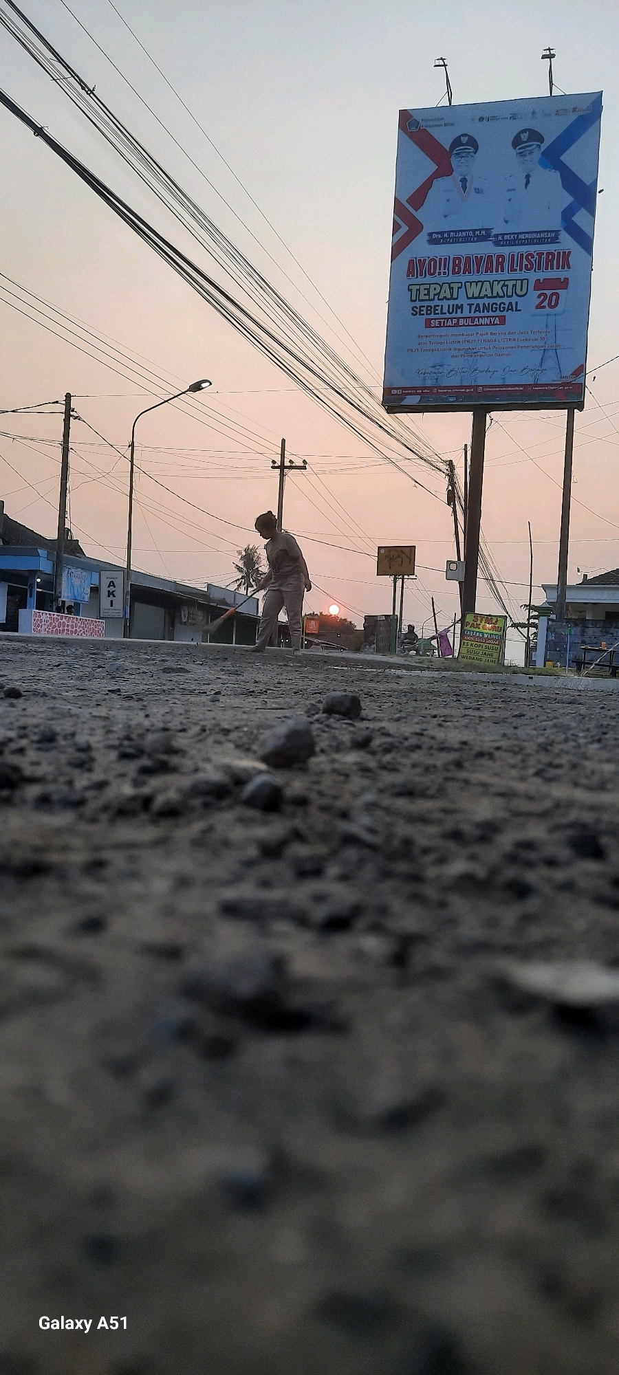 The photograph, taken from a low angle, captures a street scene at dusk in Selopuro, Indonesia. In the foreground, the rough, gravelly texture of the road fills the bottom half of the frame, with several small stones scattered across it. In the middle ground, a person wearing a light-colored uniform, possibly a worker, is sweeping the street with a broom. Their back is to the camera, and they are positioned in front of a row of buildings. To the right, a small, bright orange sun is visible near the horizon. Above the street, a dense network of power lines crisscrosses the sky, and a tall billboard advertising electricity bill payment is prominently displayed. The billboard features images of two men in formal attire and text in Indonesian. Behind the sweeping individual, streetlights and utility poles are also visible. The overall atmosphere suggests a typical evening in a semi-urban Indonesian setting.
