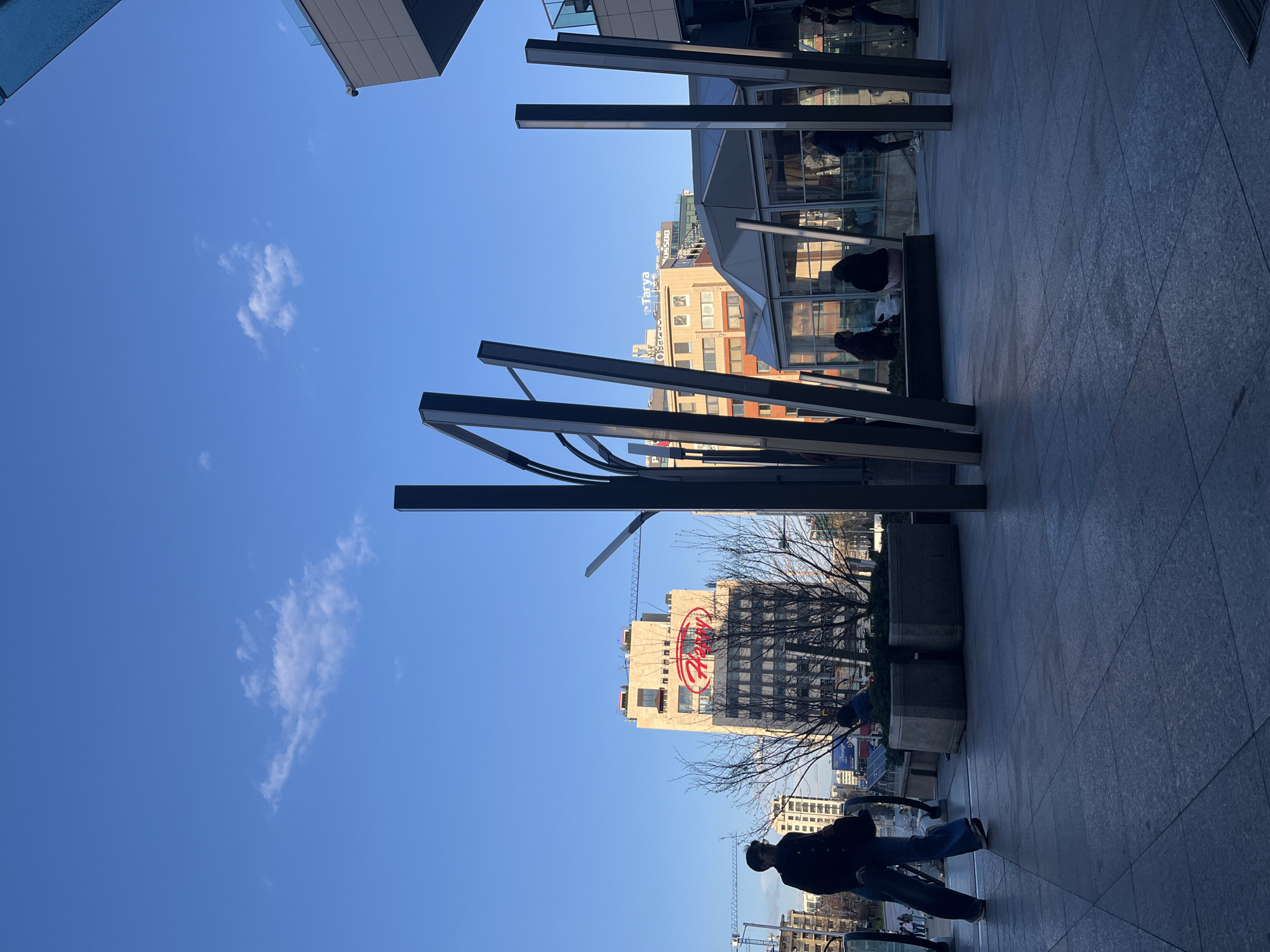 The image captures a street scene in Sofia, Bulgaria. It is oriented vertically, with the sky dominating the upper left portion. The sky is a clear blue with scattered wisps of white clouds. Modern architecture frames the shot, with building structures on both sides. A series of black metal beams create a canopy-like structure overhead, adding a geometric element to the composition. Further down the street, the distinctive "Harveys" logo is visible on a building. In the foreground, a person wearing jeans and a black coat walks away from the viewer. A bicycle or scooter is visible near the person. The ground is paved with gray stone tiles, reflecting the light. Other people are visible under a covered area, likely seated. The scene is captured during daylight hours, with the sun casting shadows on the ground and buildings.