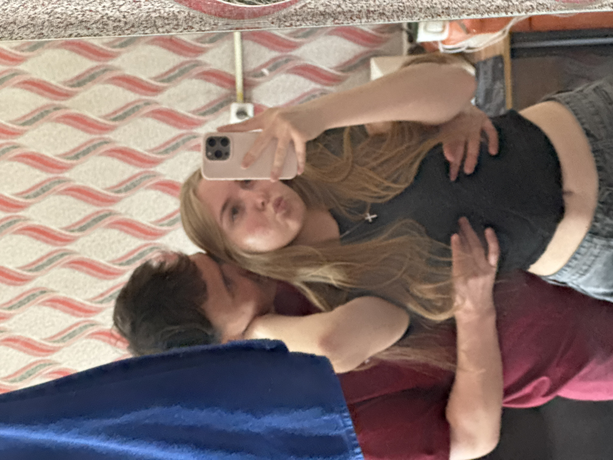 A selfie is taken in a mirror showing a couple, presumably in Perm, Russia. The woman is holding a phone with her left hand, puckering her lips for the picture. She has long blonde hair, a dark top, and gray denim bottoms. The man has dark hair and is wearing a maroon shirt, embracing her from behind. The background reveals a wavy red and white patterned wall and a possible vanity or storage unit. Part of a blue object, possibly a blanket or clothing item, obscures the lower portion of the image.