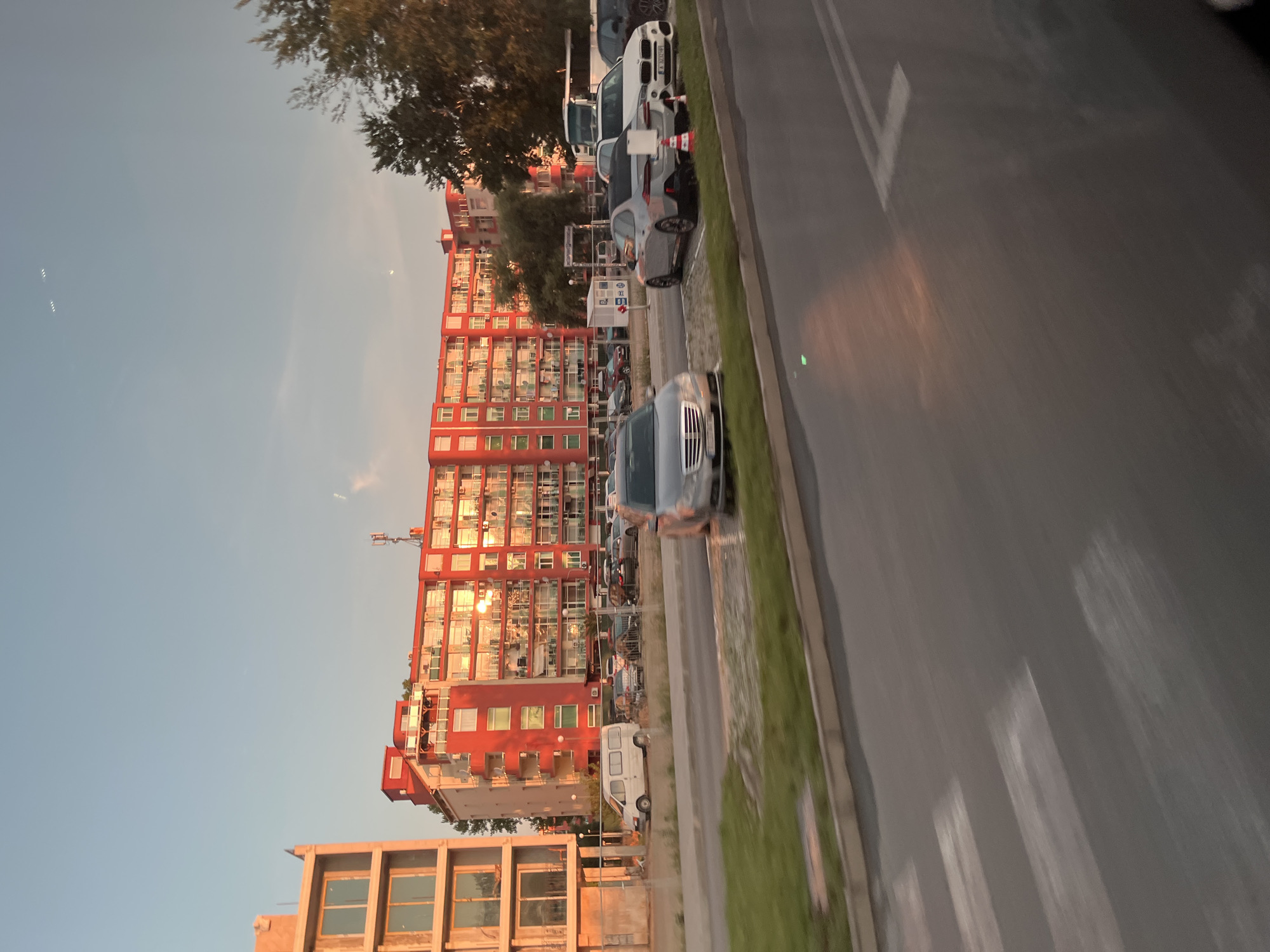 The image shows a street scene in Nesebar, Bulgaria, during sunset. A bright red apartment building with balconies is the dominant feature, reflecting the warm light of the setting sun. In front of the building, several cars are parked, including a white van and a silver sedan. A construction site with a fence is visible to the left of the building, with an unfinished building in the foreground. The street in front of the parking lot has a crosswalk. Trees line the right side of the image, and the sky is a clear blue with a few wisps of clouds. The overall impression is of a residential area in a coastal town during a pleasant evening.