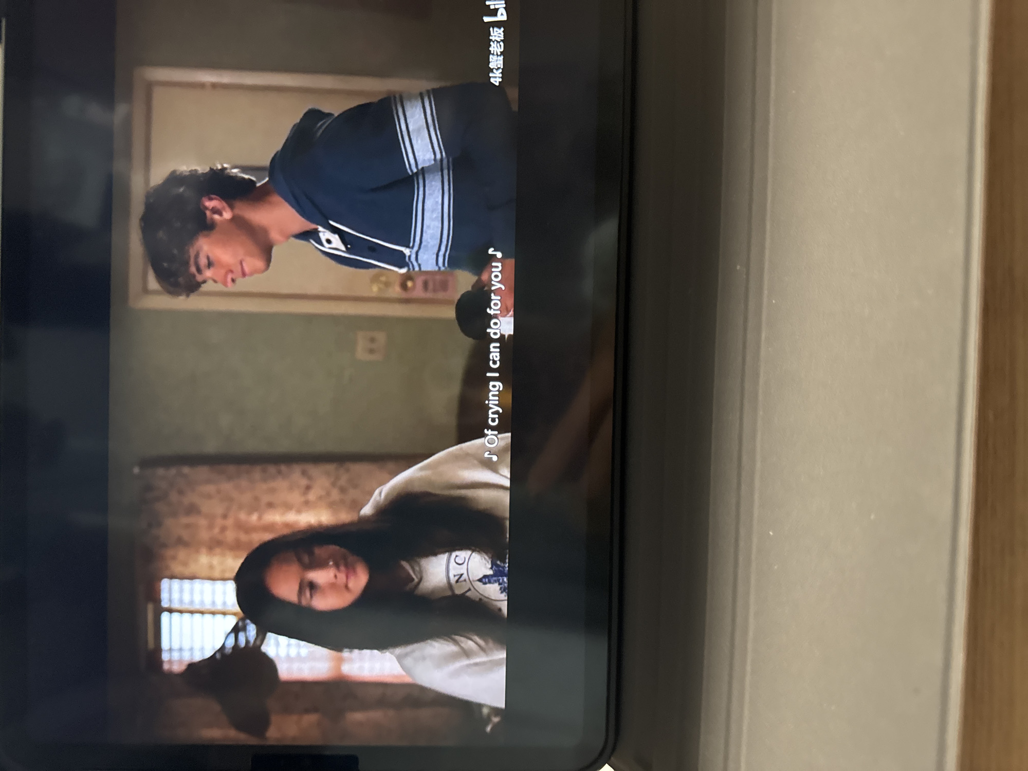 A tablet screen displays a scene with two actors. A male actor is shown from the chest up, wearing a dark blue sweatshirt with white stripes. Below him is a female actor, also from the chest up, wearing a white sweatshirt with a logo on the front. Subtitles in white are present on the screen. The surrounding environment shows the edge of a tablet and a wooden surface. There is no indication of the location being in Nan'an District, China within the image itself.
