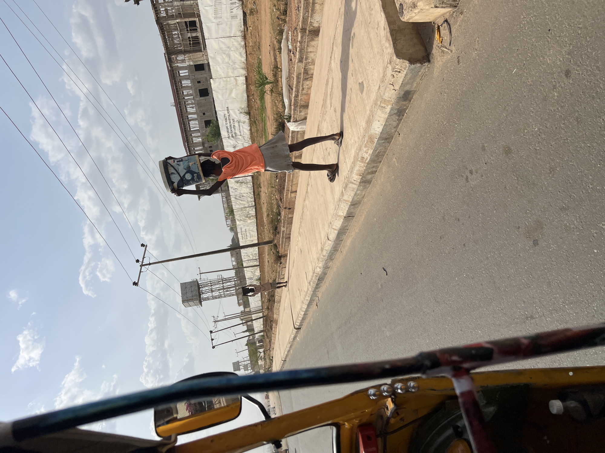 A photograph shows a man in an orange shirt and gray skirt walking along a sidewalk in Minna, Nigeria. The man is carrying a heavy box overhead. The sidewalk is next to a road. A vehicle, possibly an auto rickshaw, is visible in the foreground. Behind the man, there is an under-construction building and an electricity pole. The sky is bright with scattered clouds.