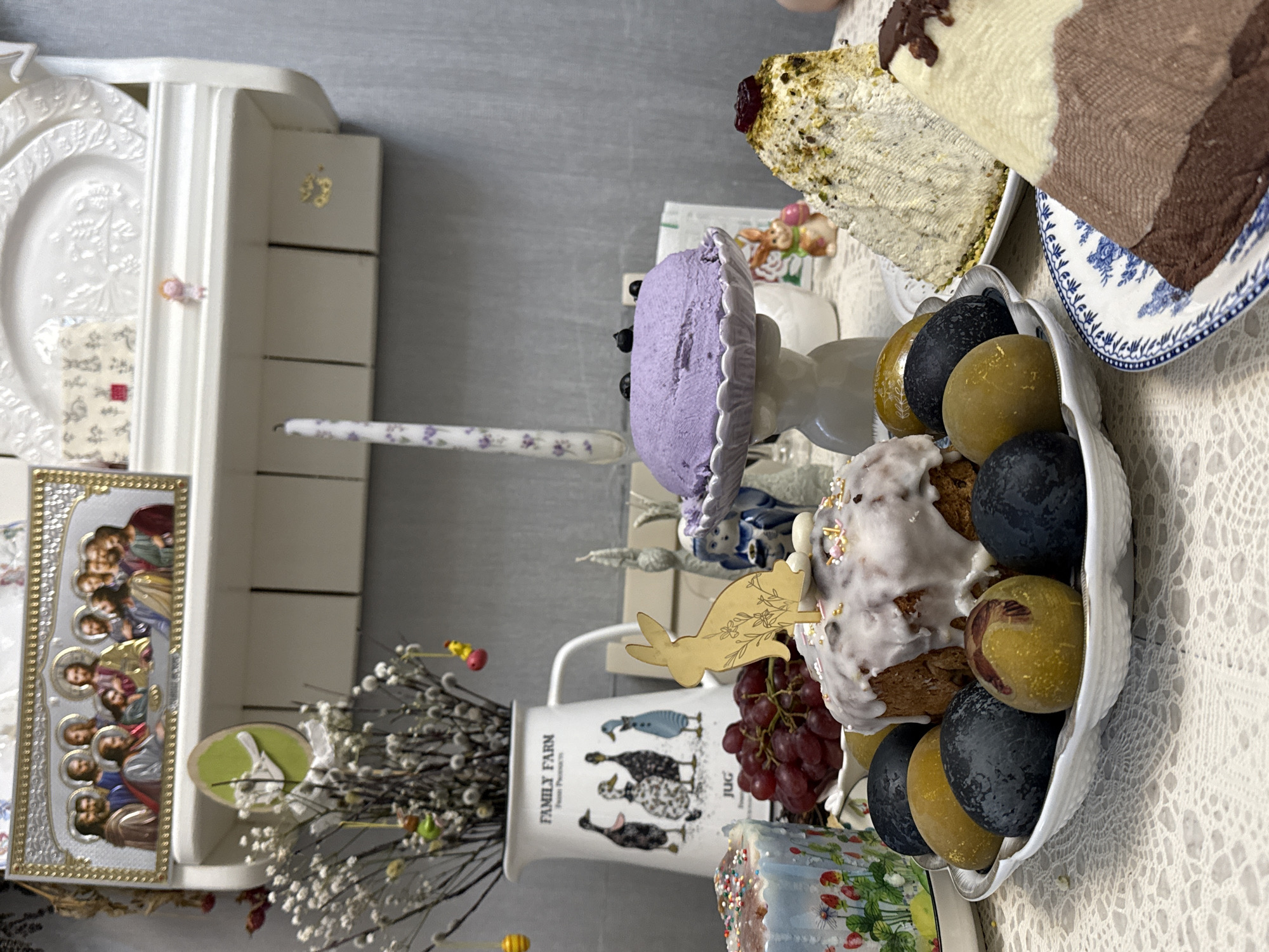 A tabletop arrangement shows Easter-themed food and decorations. There are several baked goods, including what appears to be kulich bread with white icing and decorative sprinkles, alongside colored Easter eggs in a white, shell-shaped dish. Also present are several other cakes, grapes, and a pitcher decorated with ducks. A framed religious icon is situated near a vase filled with blossoming branches, likely pussy willow. The setting appears to be inside, possibly in a home in Chaikovsky, Russia, given the style of the icon and the Easter food, which are common traditions in that region. The overall presentation suggests a celebratory Easter display.